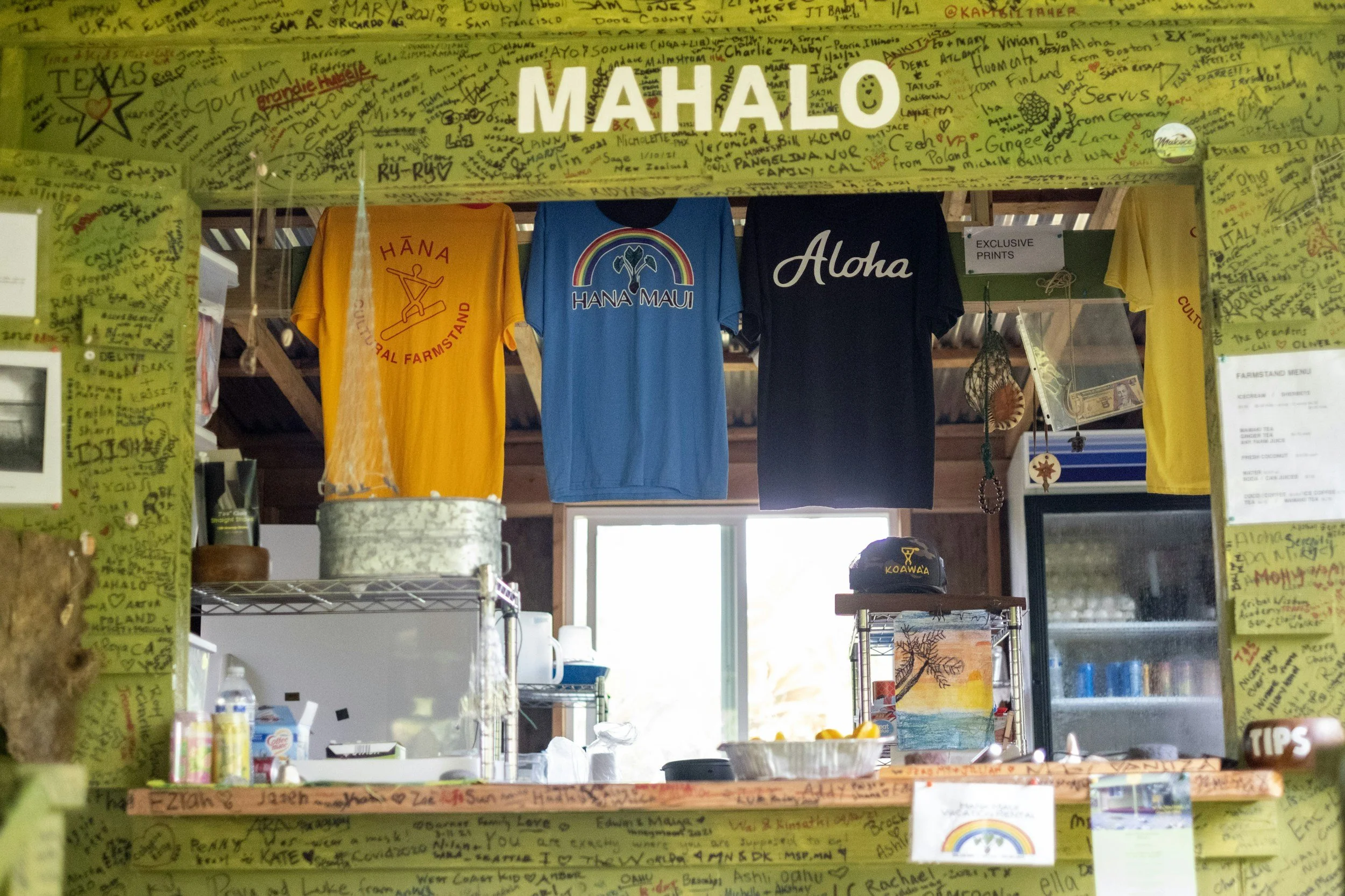 The image shows three T-shirts hanging from a horizontal bar, with the words 'HANA HANA MAUI' and 'Aloha' printed on them. The scene is set inside a shop with handwritten notes and signatures on yellow paper surrounding the display.