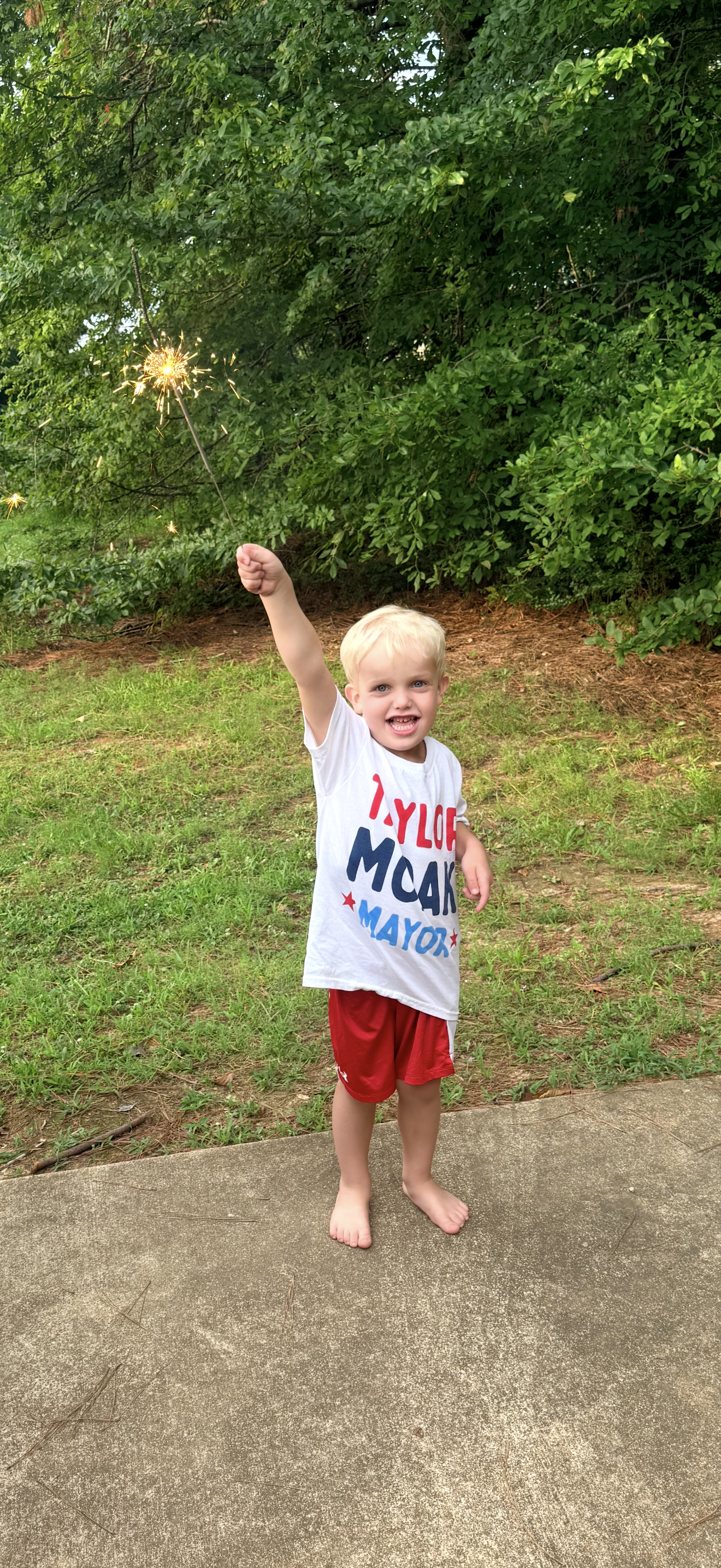 A young boy with blond hair standing on a concrete sidewalk outdoors, holding a lit sparkler and smiling, with green trees and grass in the background.