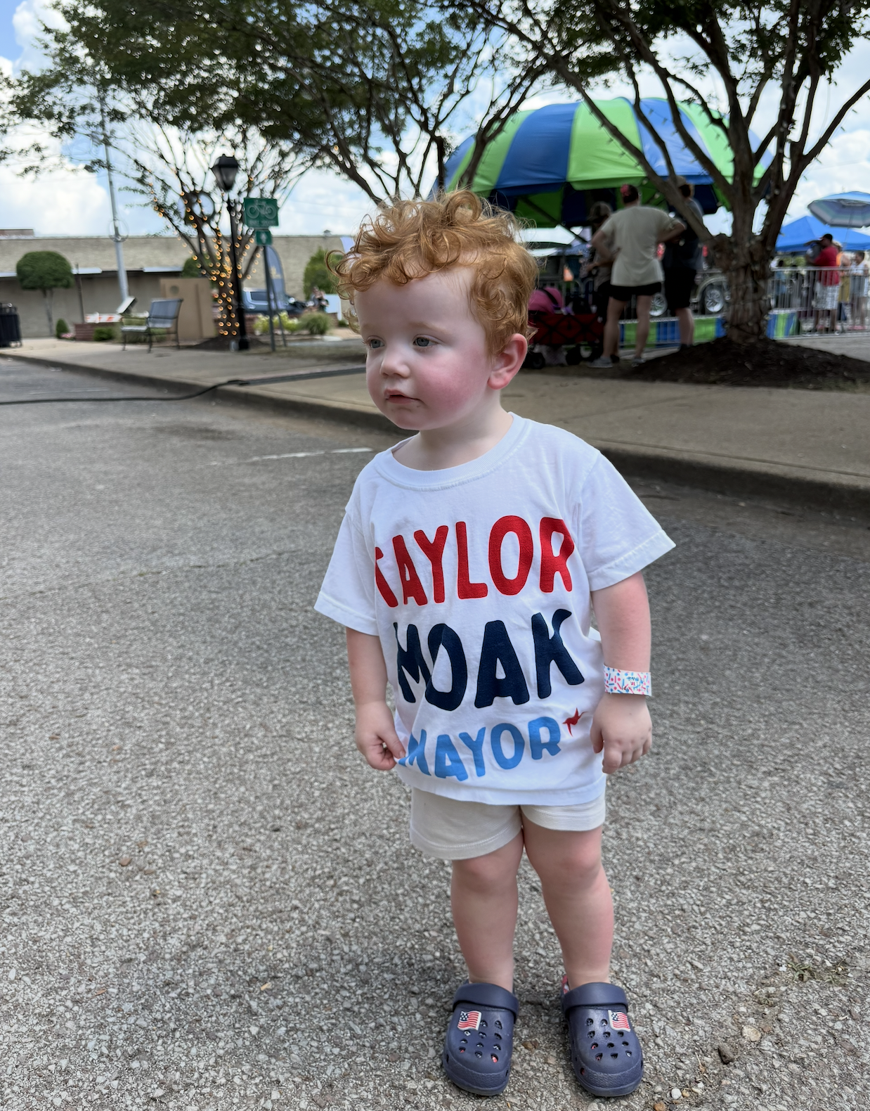 A young boy with curly red hair and fair skin standing on a paved area outdoors, wearing a white t-shirt with red, blue, and black text supporting a political candidate, beige shorts, and navy slip-on shoes with American flag design. In the backgroun