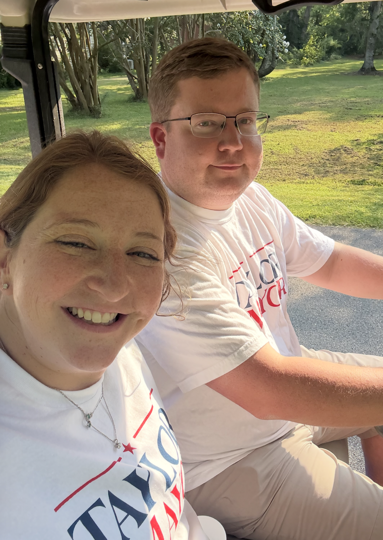 A smiling woman with freckles and red hair taking a selfie, and a man with glasses sitting next to her, both wearing white T-shirts with red and blue text, outdoors on a sunny day with a grassy and wooded background.