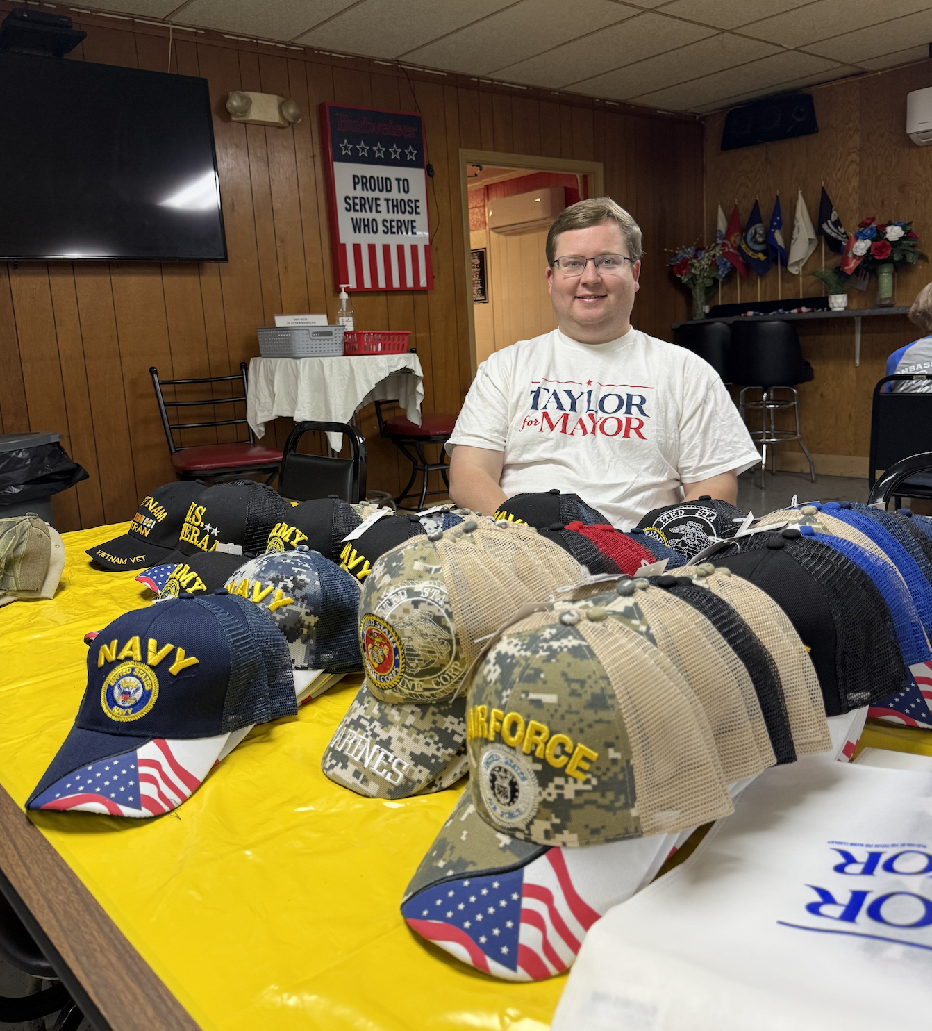 A man sitting at a table displaying various military-themed caps, including Navy, Army, and Air Force hats. The man is wearing a white T-shirt that says 'TAYLOR for MAYOR.' The background shows wood-paneled walls, a TV, flags, and patriotic decoratio