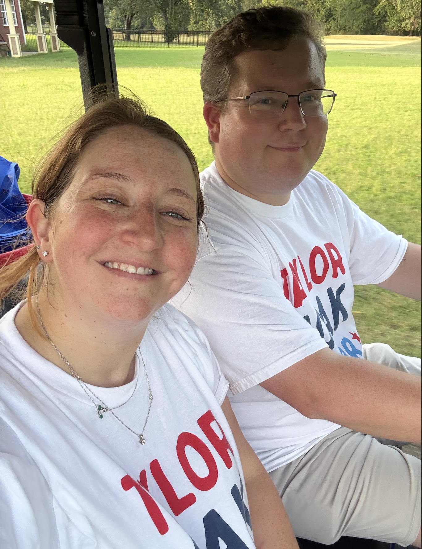Two smiling people riding in a golf cart on a grassy course, both wearing white T-shirts with red and blue text, with trees and a fence in the background.
