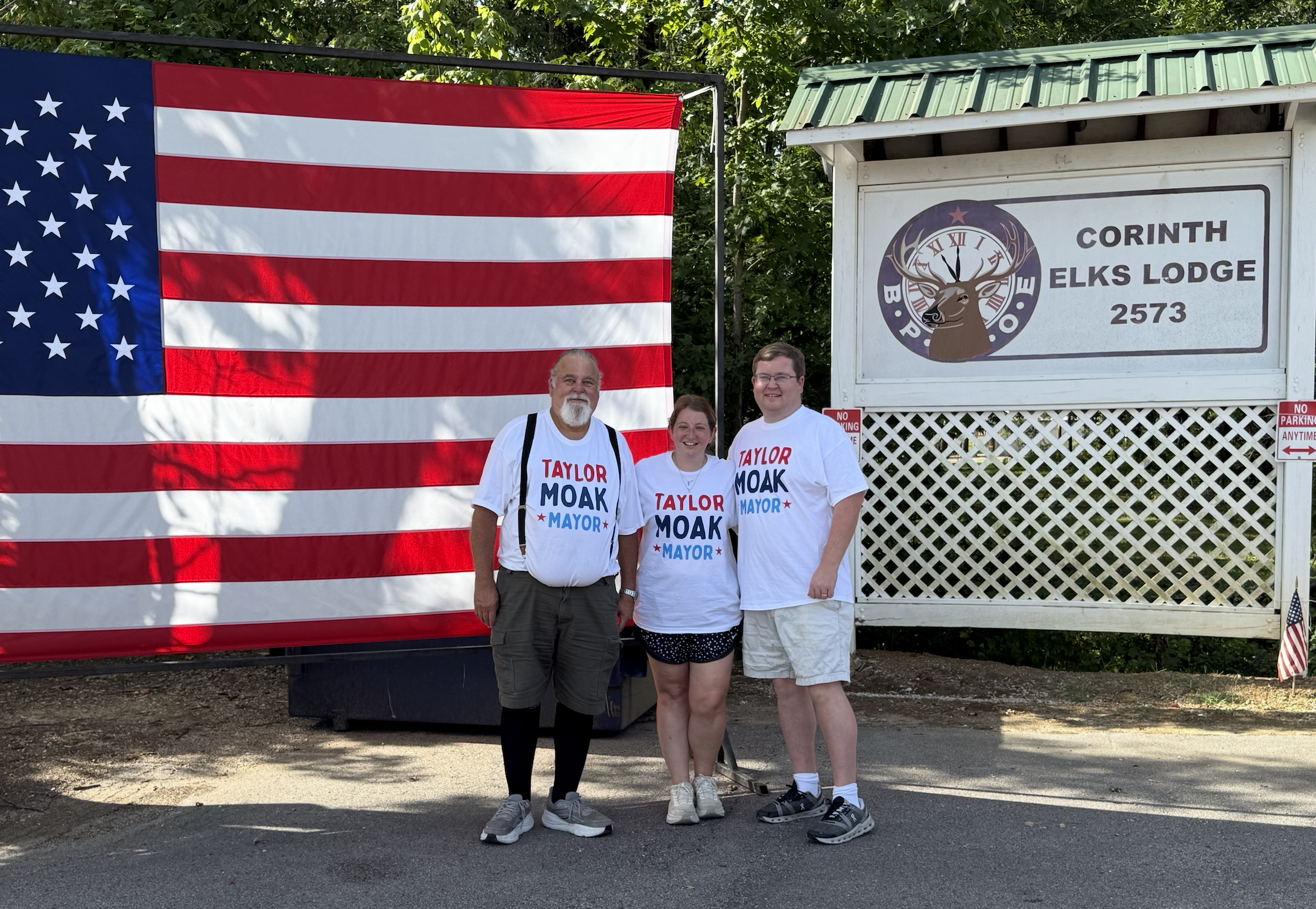 Three people standing outdoors in front of an American flag and a sign for Corinth Elks Lodge, wearing white t-shirts supporting Taylor Moak for mayor, smiling at the camera.