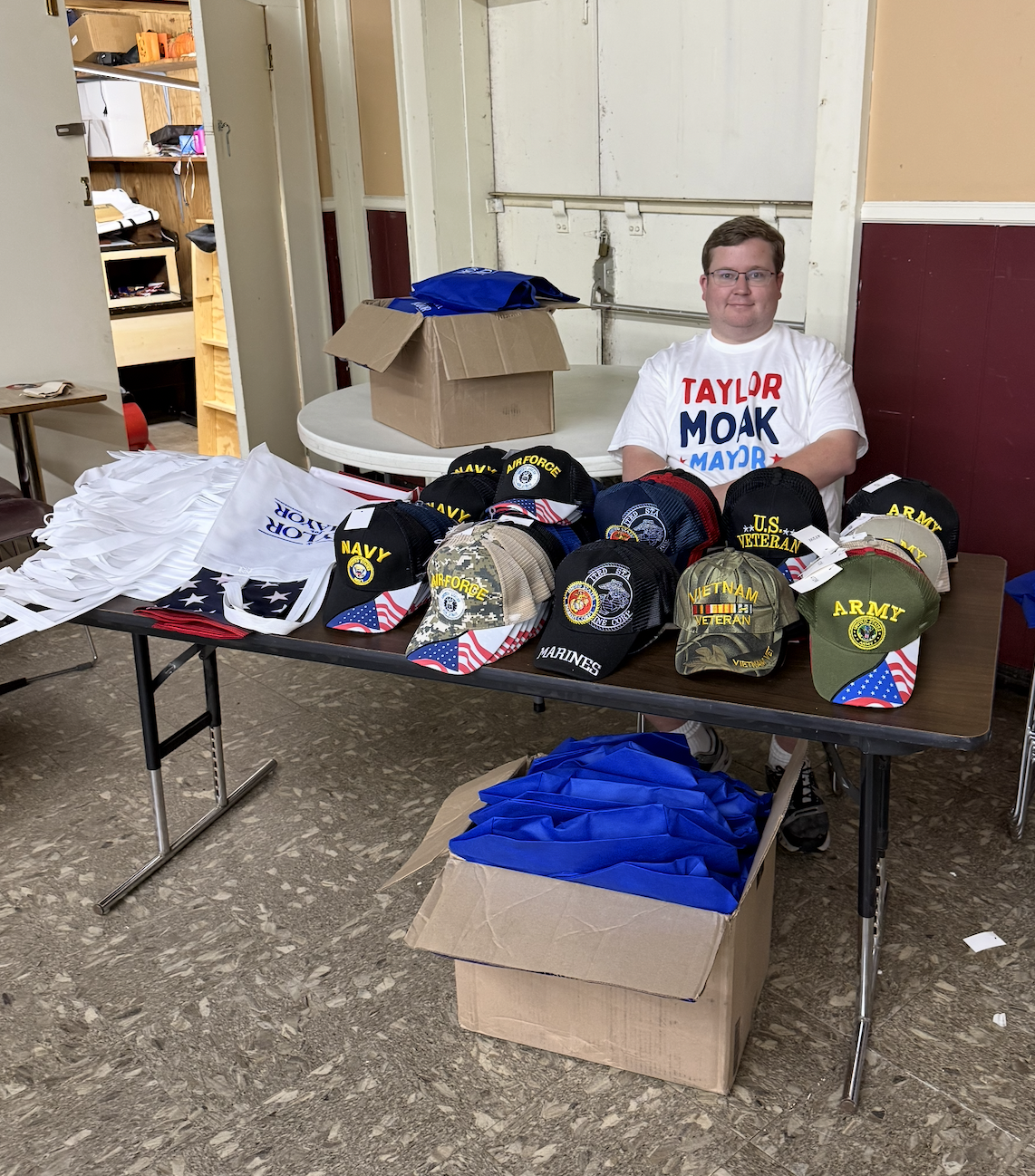 A man sitting behind a table displaying various hats with U.S. military and veteran logos, including Navy, Army, Air Force, Marine, U.S. Veteran, and Vietnam Veteran, inside a community hall