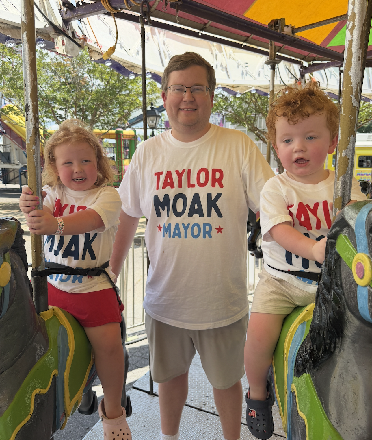 A man and two children riding on a colorful carousel at an amusement park. They are smiling and wearing T-shirts with the text "Taylor Moak Mayor." The carousel horses are decorated and the background shows trees and amusement park rides.