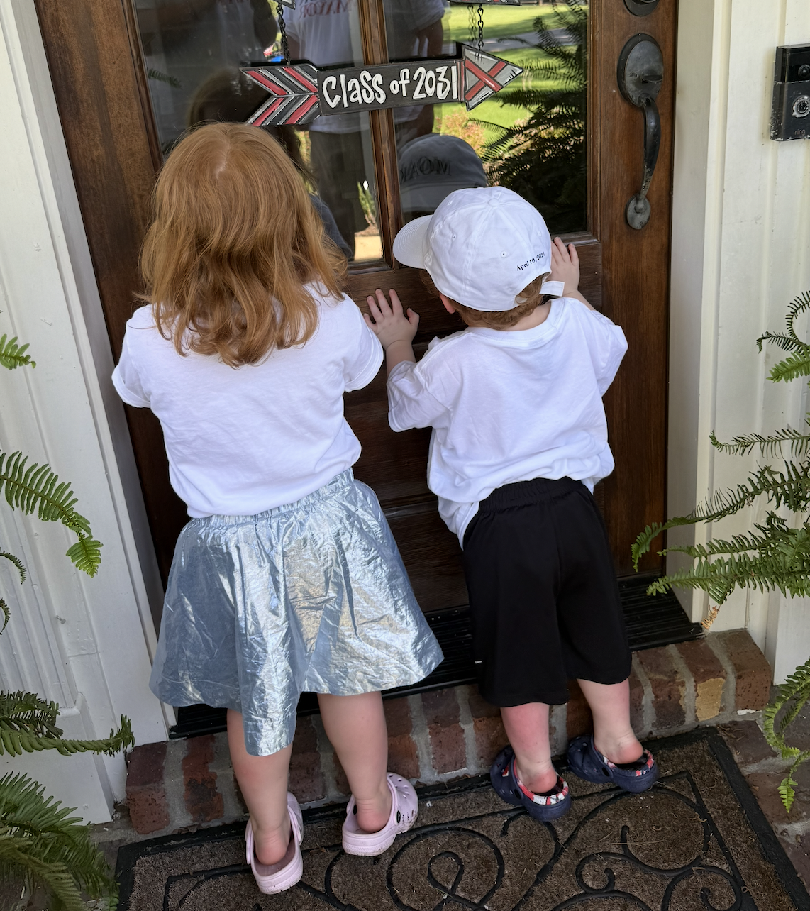 Two children, a girl with red hair and a boy with brown hair wearing a white cap, are standing on a doorstep looking through a glass door. The door has a sign that reads 'Class of 2031' hanging above it. Both children are dressed in white tops and da