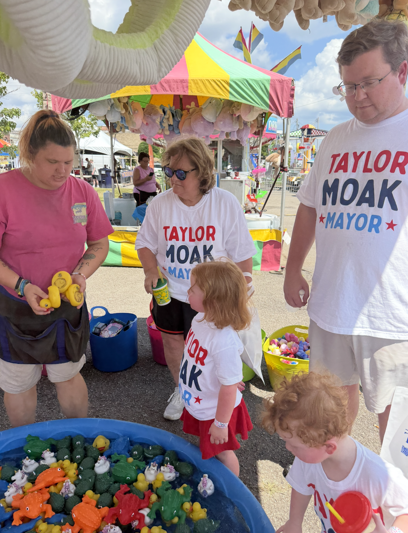 People gathered at a fair booth selling small rubber ducks and other toys, with a colorful striped tent, cotton candy, and carnival rides in the background.