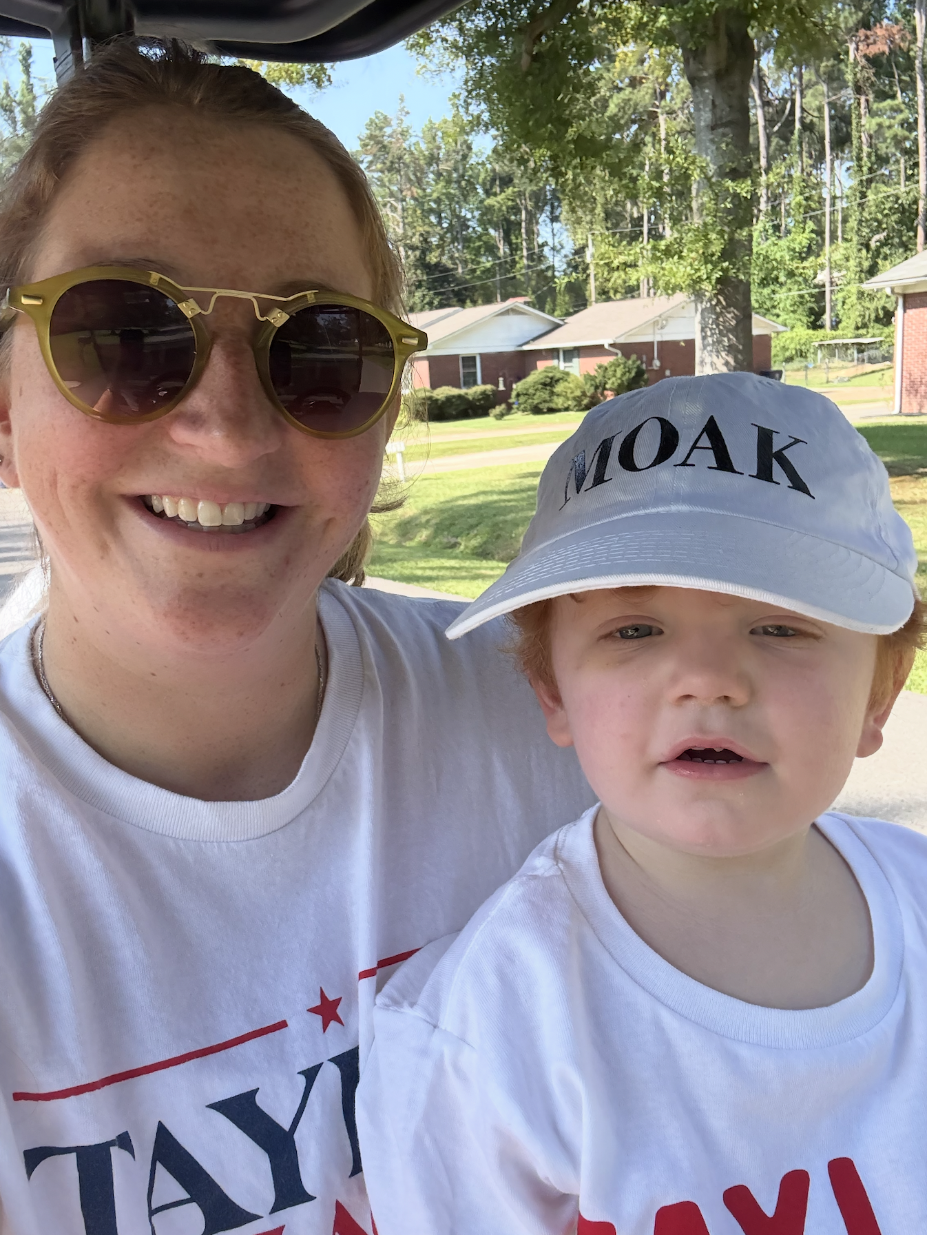A smiling woman with red hair, sunglasses, and freckles, taking a selfie with a young boy wearing a white cap and a white T-shirt, outdoors with trees and houses in the background.