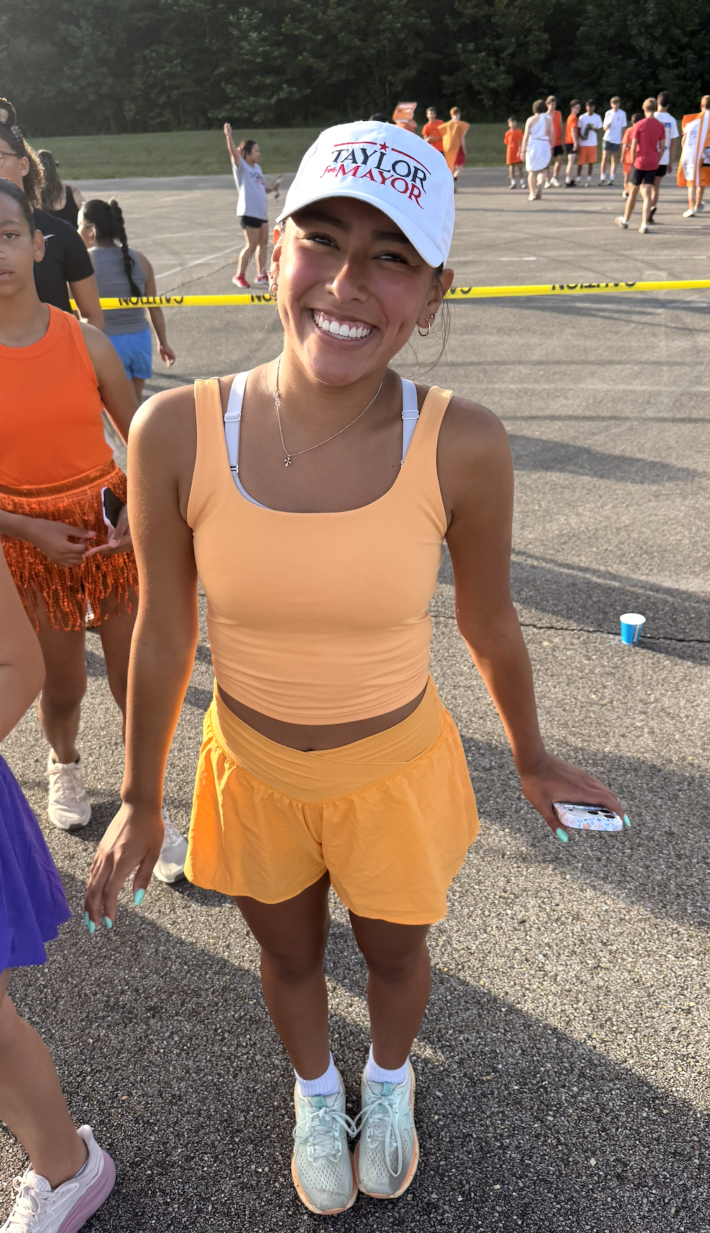 A smiling young woman wearing a white campaign hat that says "Taylor for Mayor," a peach workout top, matching orange shorts, and sneakers, standing outdoors with a group of people participating in an event or race, with yellow caution tape in the ba