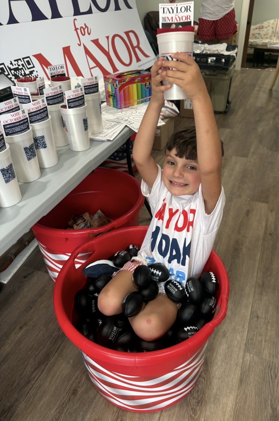 A young boy sitting in a large red and white striped bucket filled with black foam footballs, holding one raised above his head, smiling happily. The boy is wearing campaign-themed clothing for Taylor for Mayor. Nearby, there are campaign signs, butt