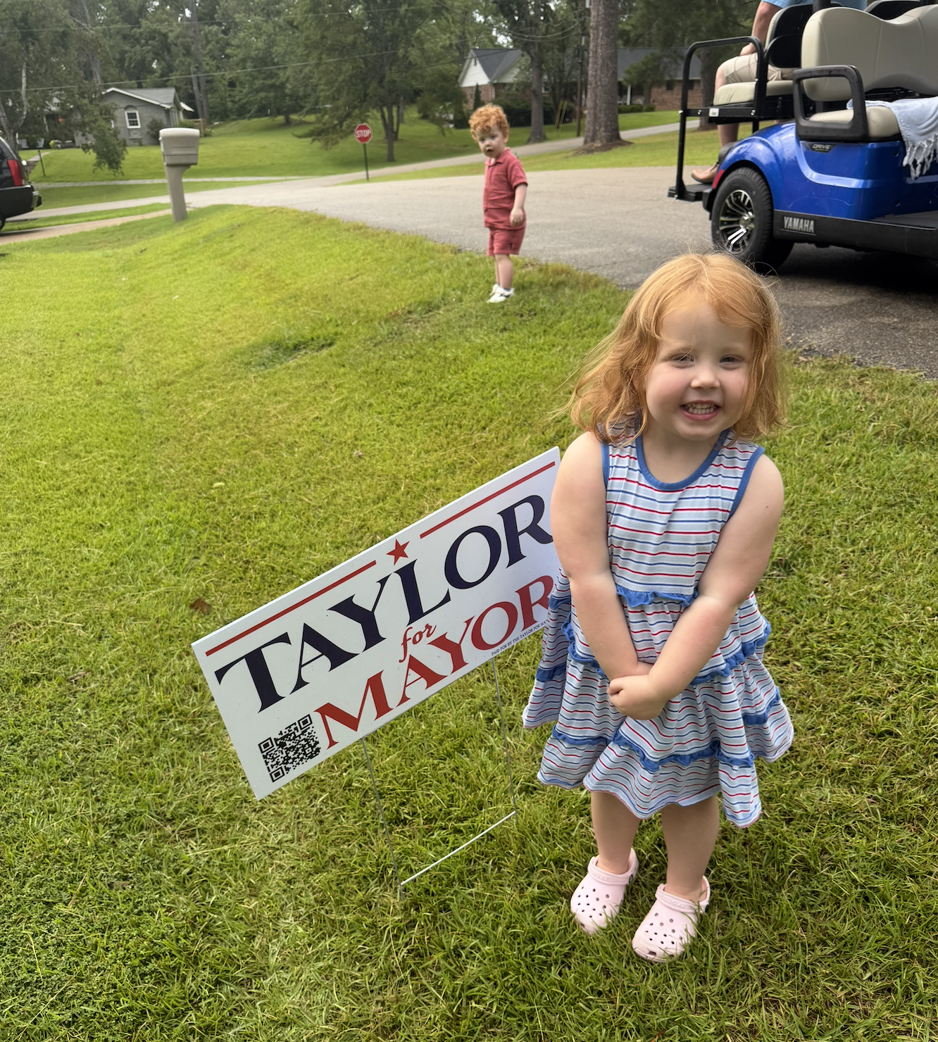 A young girl with red hair and a striped dress standing on grass, smiling at the camera, near a political campaign sign that says "TAYLOR for MAYOR" with a QR code. In the background, a boy with curly hair in a red outfit is walking near a driveway, 
