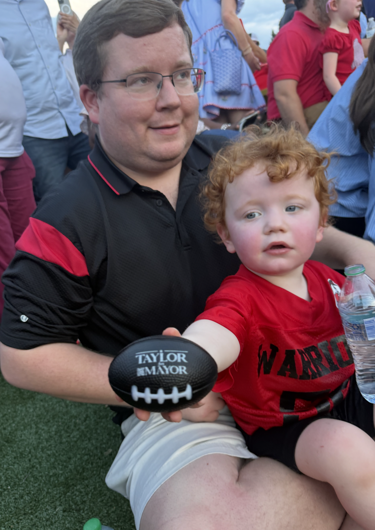 A man wearing glasses and a black and red shirt is sitting on the grass, holding a small football with 'TAYLOR MAYOR' written on it. A young red-haired boy in a red shirt is sitting on his lap, holding a water bottle, with a slightly serious expressi