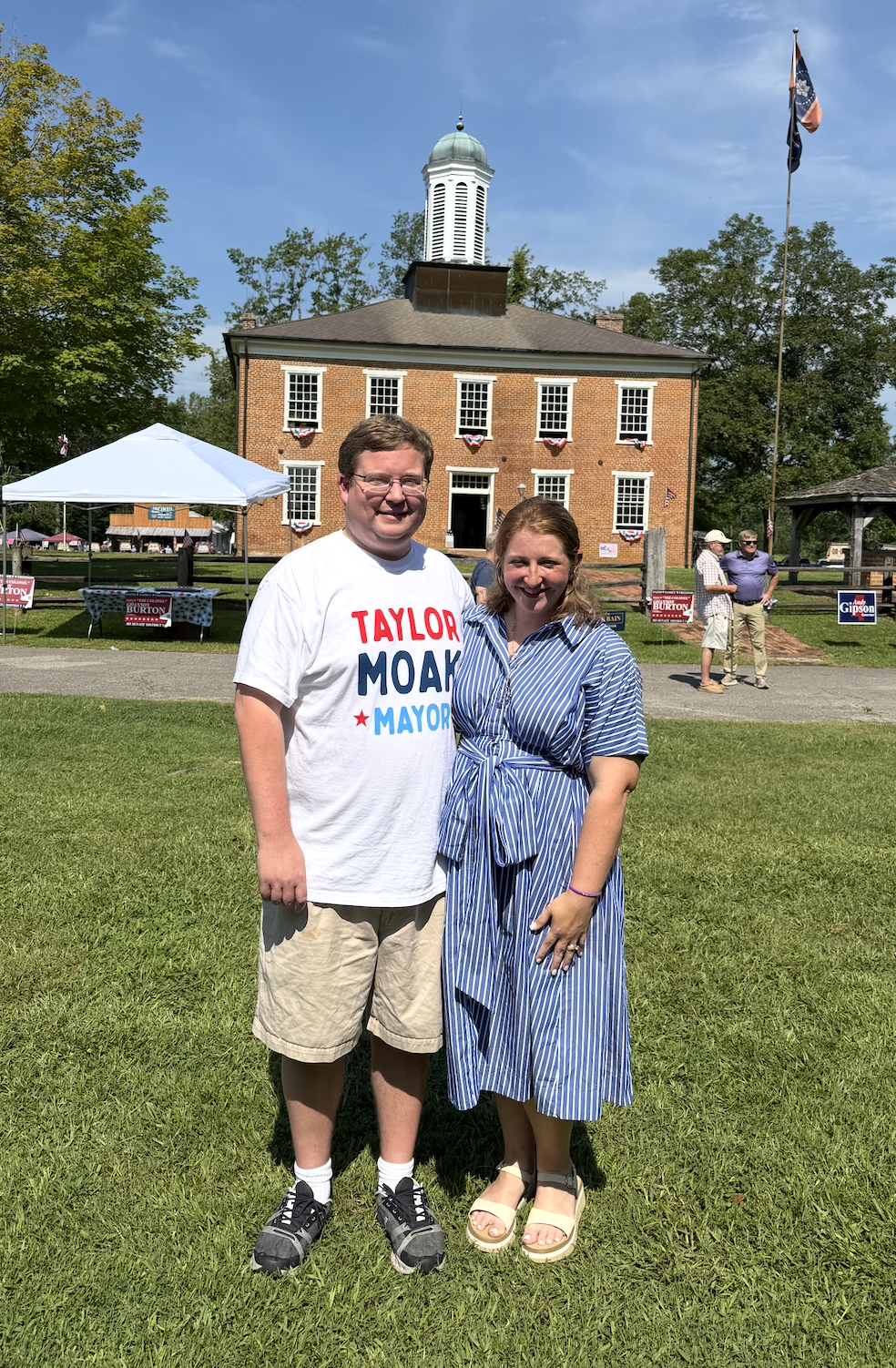 A man and a woman standing on grass in front of a historic brick building with a white cupola, during a sunny day. The man is wearing a white t-shirt with red and blue text supporting a mayoral candidate, and khaki shorts. The woman is wearing a blue