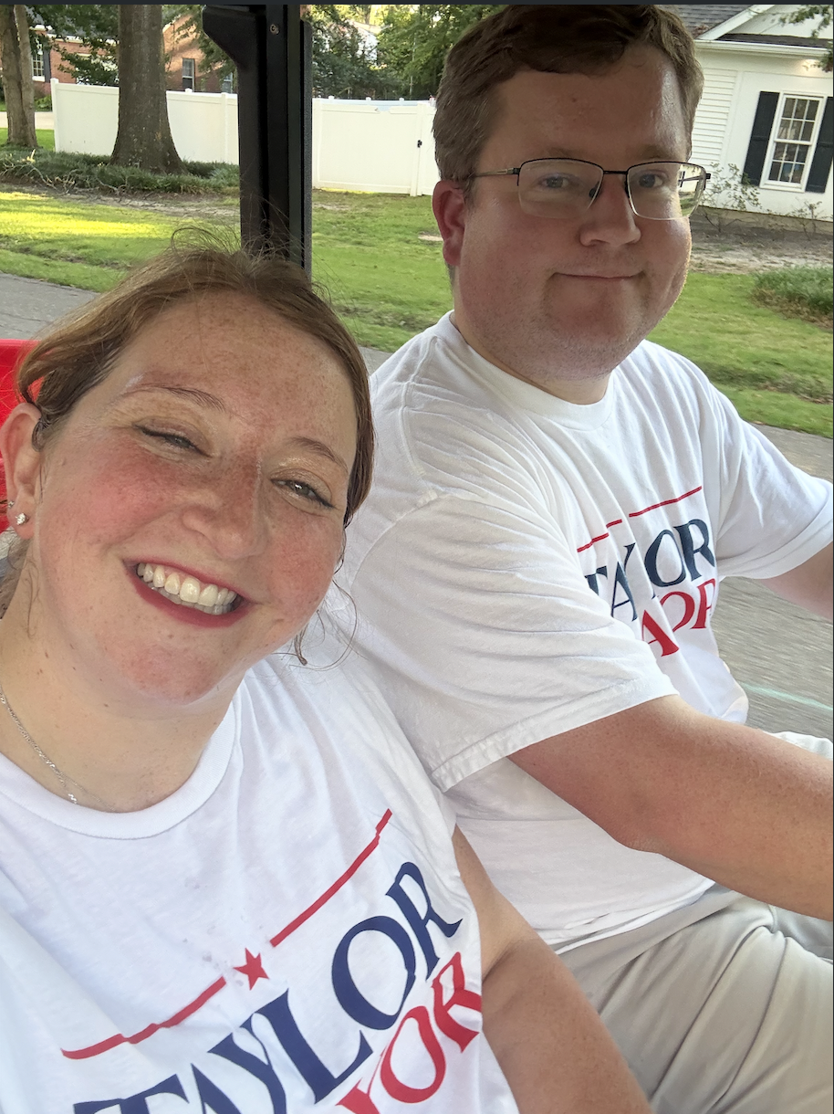 A smiling woman and a man sitting together outdoors, both wearing white T-shirts with red and blue text, on a sunny day. The background shows trees, grass, a white fence, and a white house with black shutters.