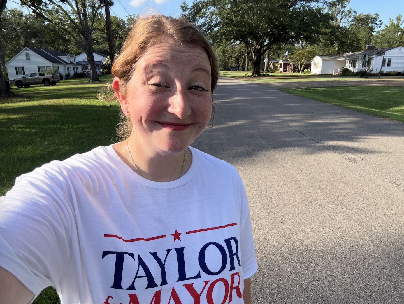 Woman taking a selfie outdoors on a sunny day, wearing a white T-shirt with the text "TAYLOR for MAYOR," standing on a residential street with green grass, trees, and houses in the background.