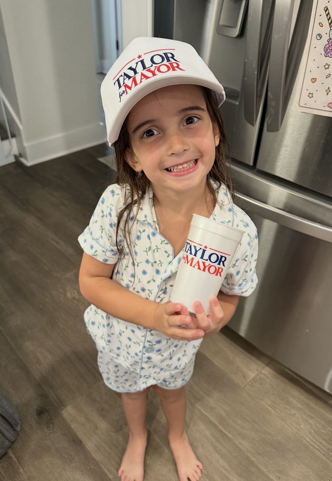 A young girl with long brown hair wearing pajamas stands indoors by a stainless steel refrigerator. She is smiling with her teeth showing and holding a large white cup. She is wearing a white cap with red and blue text and stars, reading "TAYLOR for 