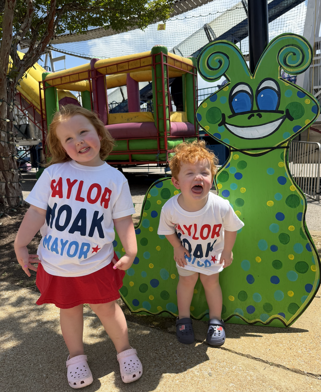 Two young children posing in front of a colorful, cartoonish butterfly cutout at an outdoor playground. The girl on the left has red hair, is smiling, and is wearing a white T-shirt with red and blue text and a red skirt, and pink Crocs. The boy on t