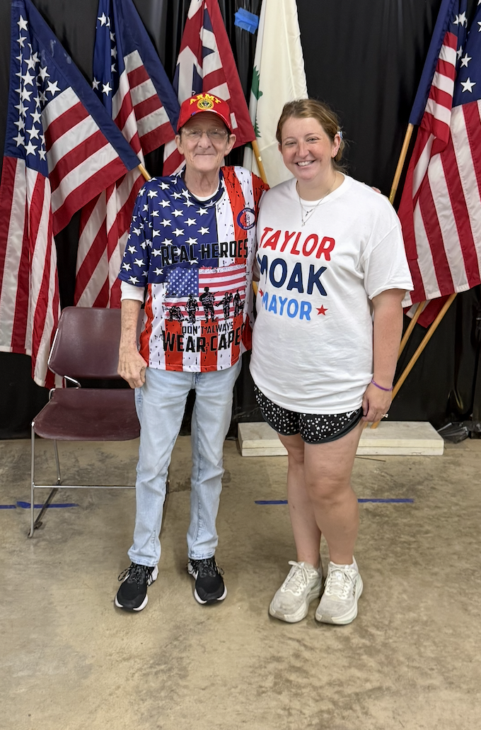 Two women standing in front of American flags, smiling. One woman is elderly, wearing a patriotic shirt and red cap. The other woman is young, wearing a white T-shirt with candidate's name and mayoral campaign.
