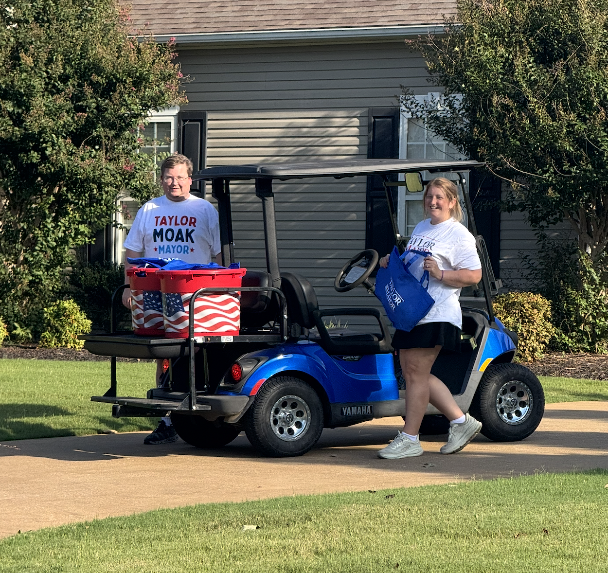 Two women wearing campaign T-shirts with the slogan 'Taylor Moak Mayor' standing and smiling near a blue golf cart, holding campaign materials, in a suburban neighborhood with trees and a house in the background.