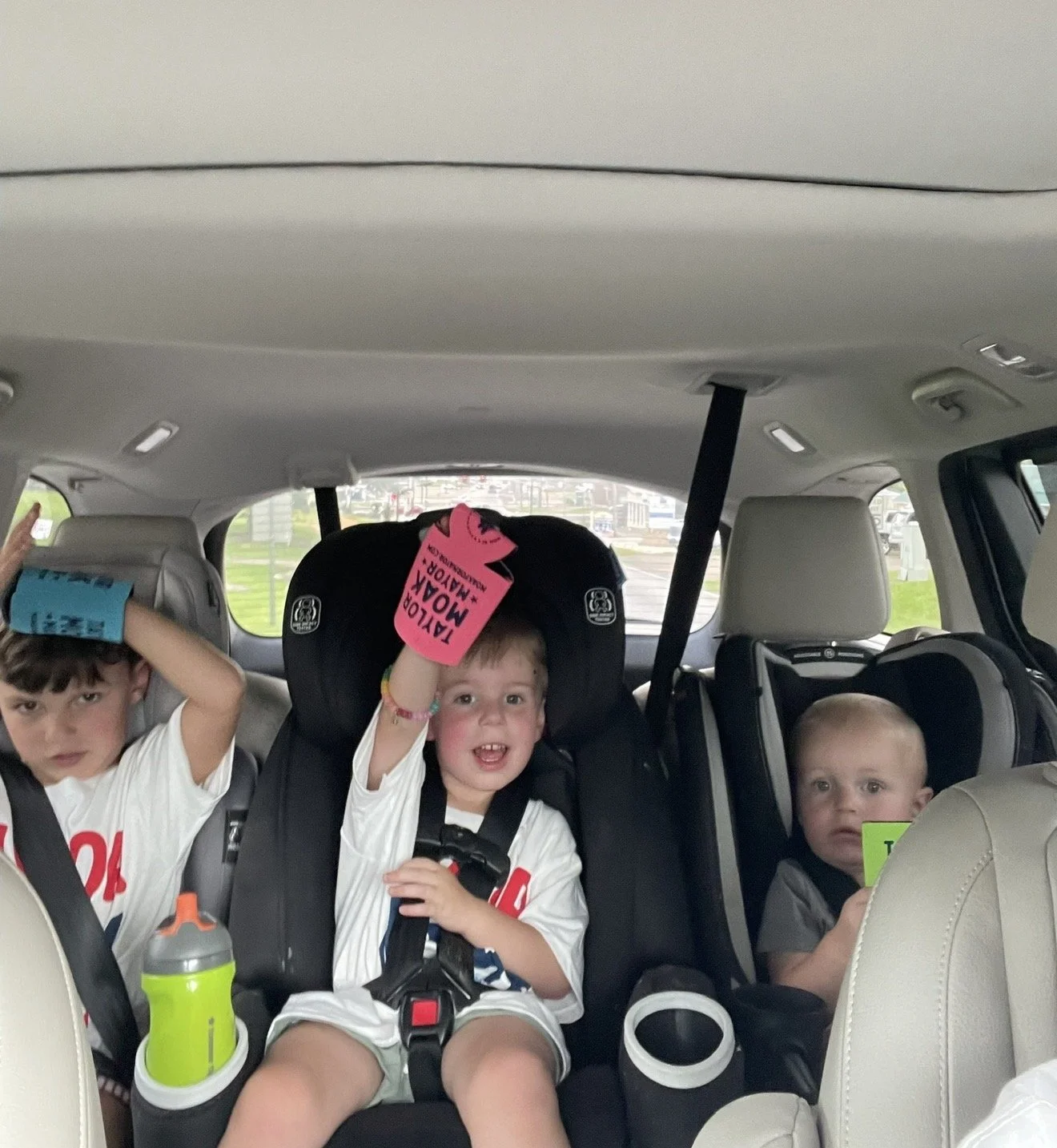 Three young boys seated in a car, with the two in the rear seats. The boy in the middle is smiling, wearing a white shirt, and has a pink wristband and a glove with a pink tag that reads "A year of mox, matador". The boy on the right has short blond 