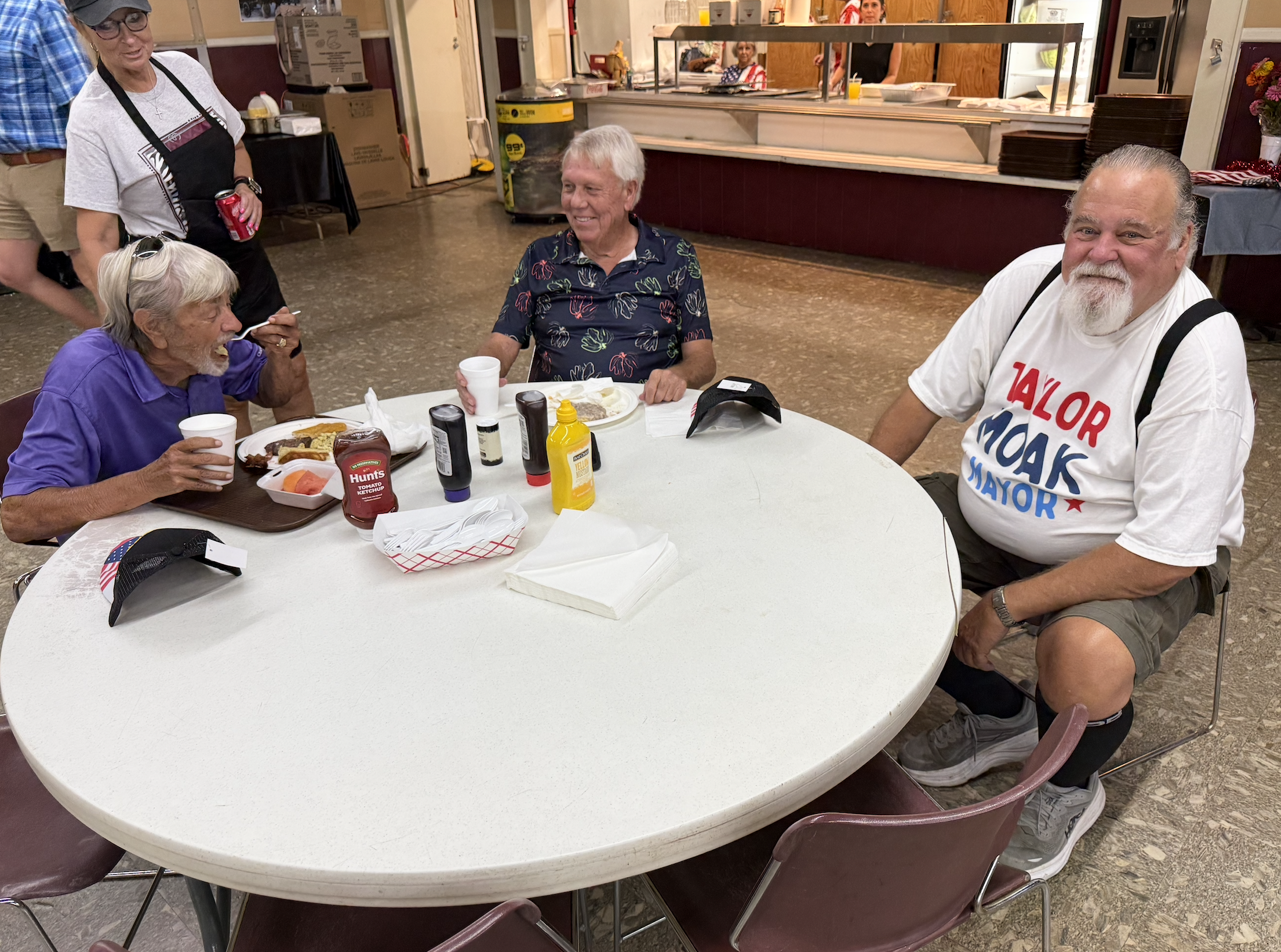 A group of five people sitting at a round table in a restaurant enjoying a meal. There are condiments including ketchup and mustard on the table, along with plates of food and cups. A woman in a white shirt and glasses is standing, while others are s