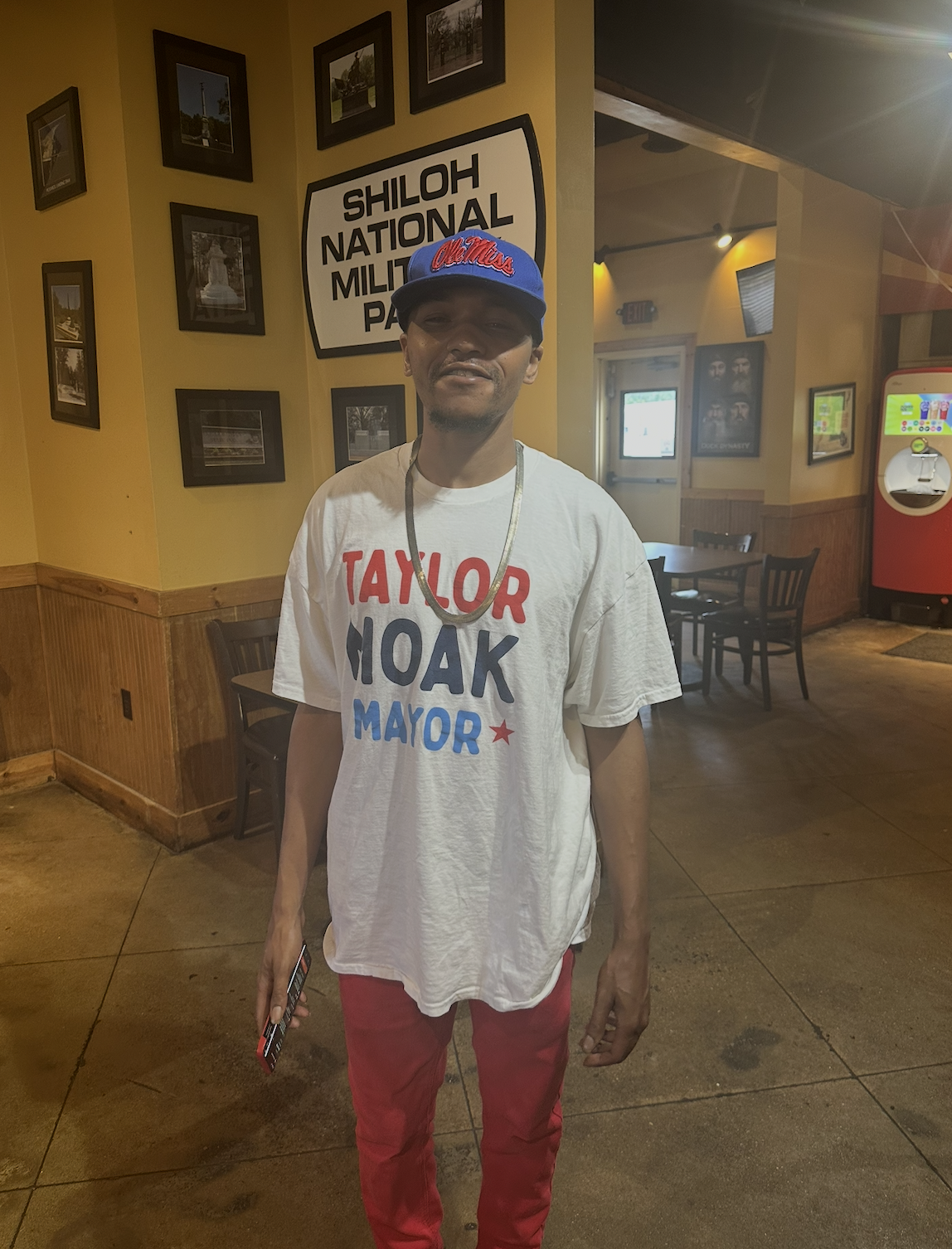 A man wearing a baseball cap and a campaign t-shirt standing inside a restaurant or cafe. The t-shirt promotes political candidates for mayor and the cap supports Ole Miss. There are framed pictures and a sign indicating Shiloh National Military Park