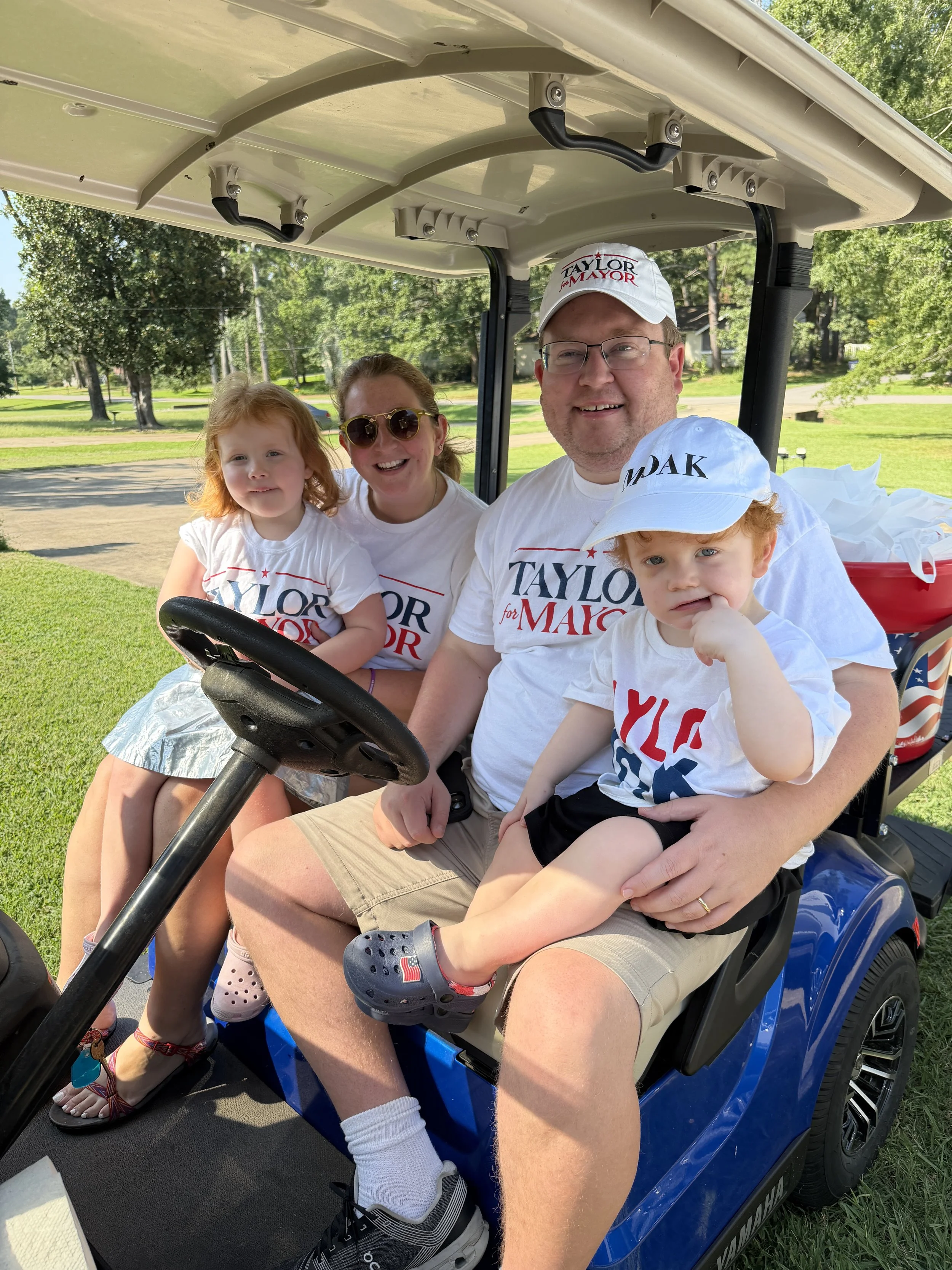 A family of four sitting in a blue golf cart outdoors on a sunny day, with green grass and trees in the background. They are all wearing white shirts that say "Taylor for Mayor". The father is wearing a white cap with the same slogan and glasses, hol