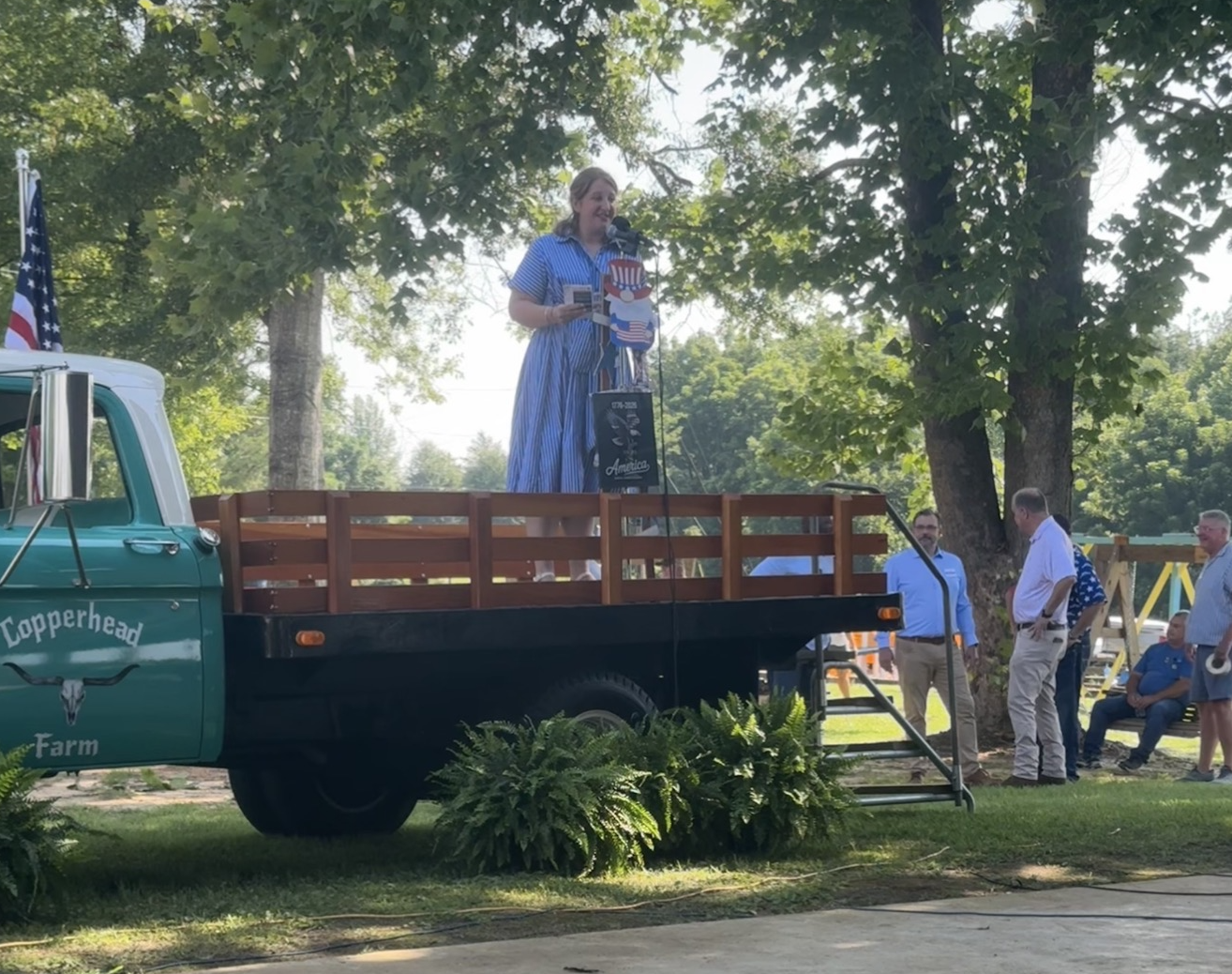 A woman dressed in blue and white striped dress standing on a flatbed truck, speaking at an outdoor event with American flags and trees in the background.