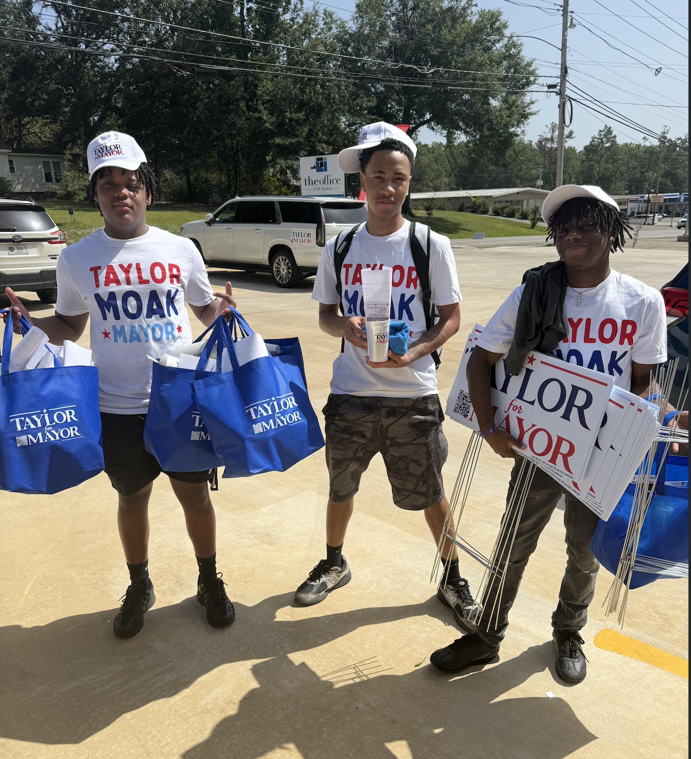 Three young men wearing 'Taylor Moak for Mayor' shirts and hats are standing outside on a sunny day, holding campaign materials and blue campaign bags. Behind them is a parking lot with cars and some trees.