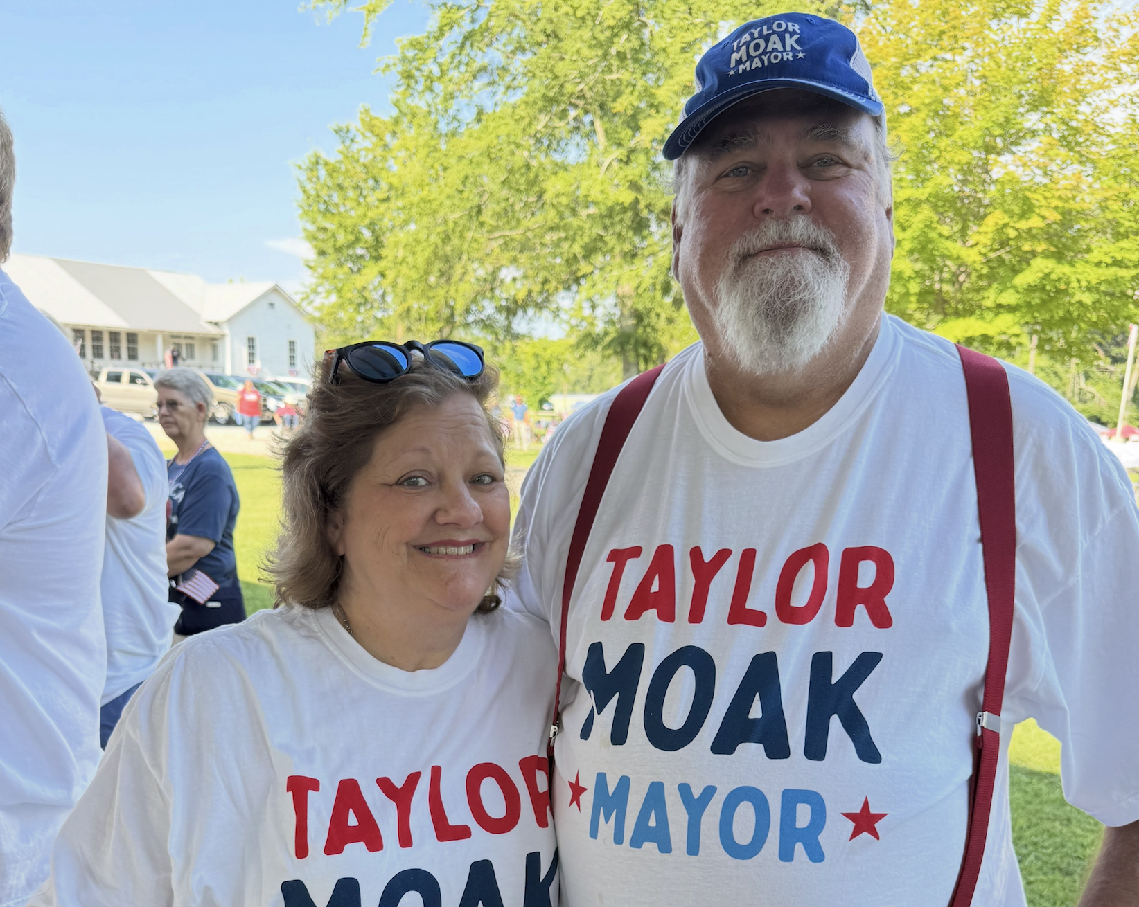 Two people at an outdoor event, both wearing white T-shirts with red and blue text supporting Taylor Moak for mayor. The man is also wearing a blue hat and has a white beard, and the woman has sunglasses resting on her head. The background features t