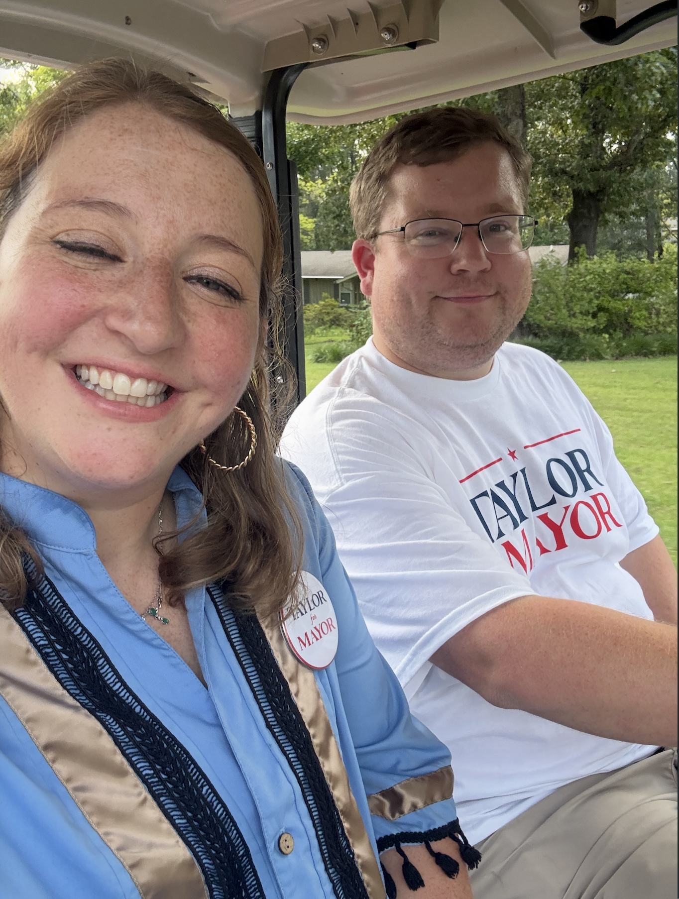 A smiling woman with light skin, freckles, and long wavy hair, wearing a blue and beige jacket with a campaign sticker that says 'TAYLOR for MAYOR,' sitting next to a man with light skin, short brown hair, glasses, and wearing a white T-shirt with th