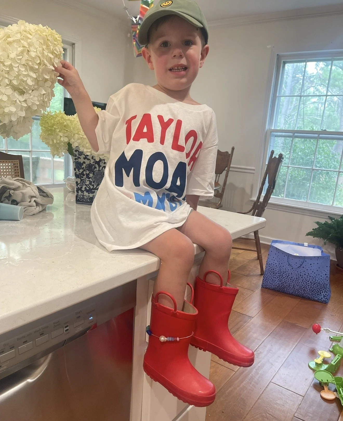 A young boy sitting on a kitchen counter, wearing red rain boots, a white t-shirt with red, blue, and black lettering, and a green cap with a smiley face. The kitchen has large windows, white walls, and various objects including a blue gift bag on th