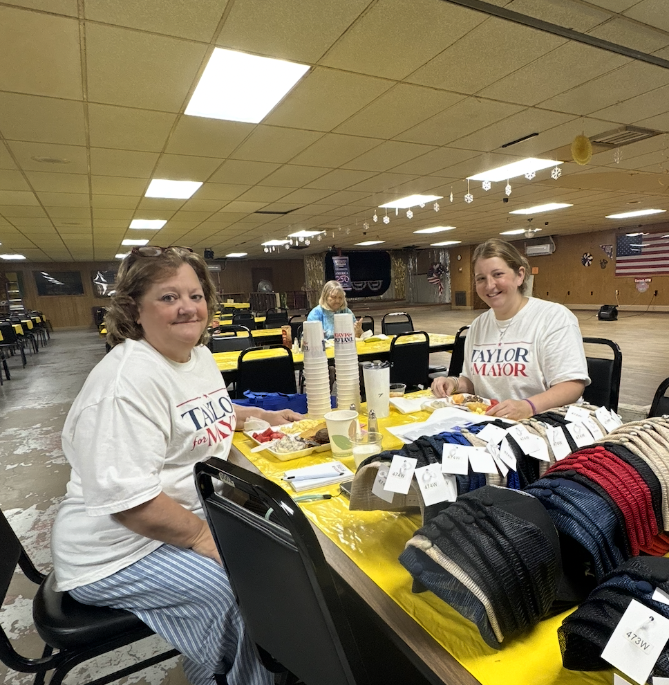 Two women sitting at a table with food and supplies in a decorated hall, wearing campaign t-shirts supporting Taylor for Mayor.