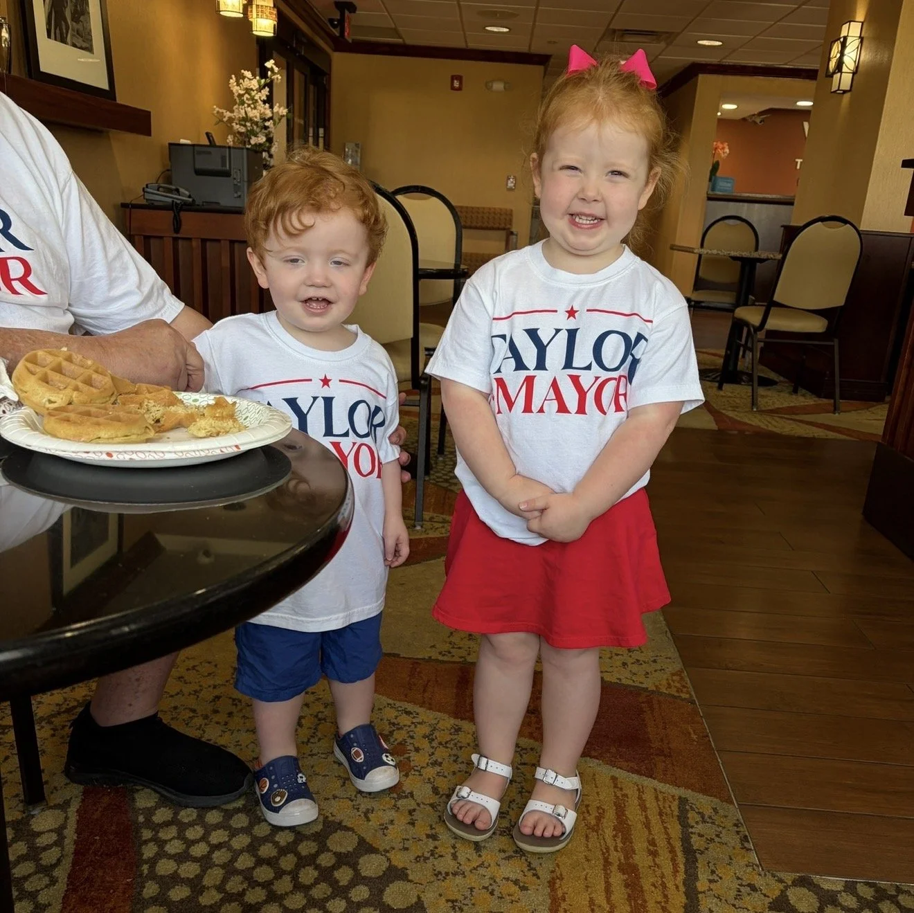 Two young children, a boy and a girl, standing in a restaurant. They are wearing matching white T-shirts with the words 'TAYLOR FOR MAYOR' printed on them. The girl has red hair tied with a pink bow and is wearing a red skirt and sandals, while the b