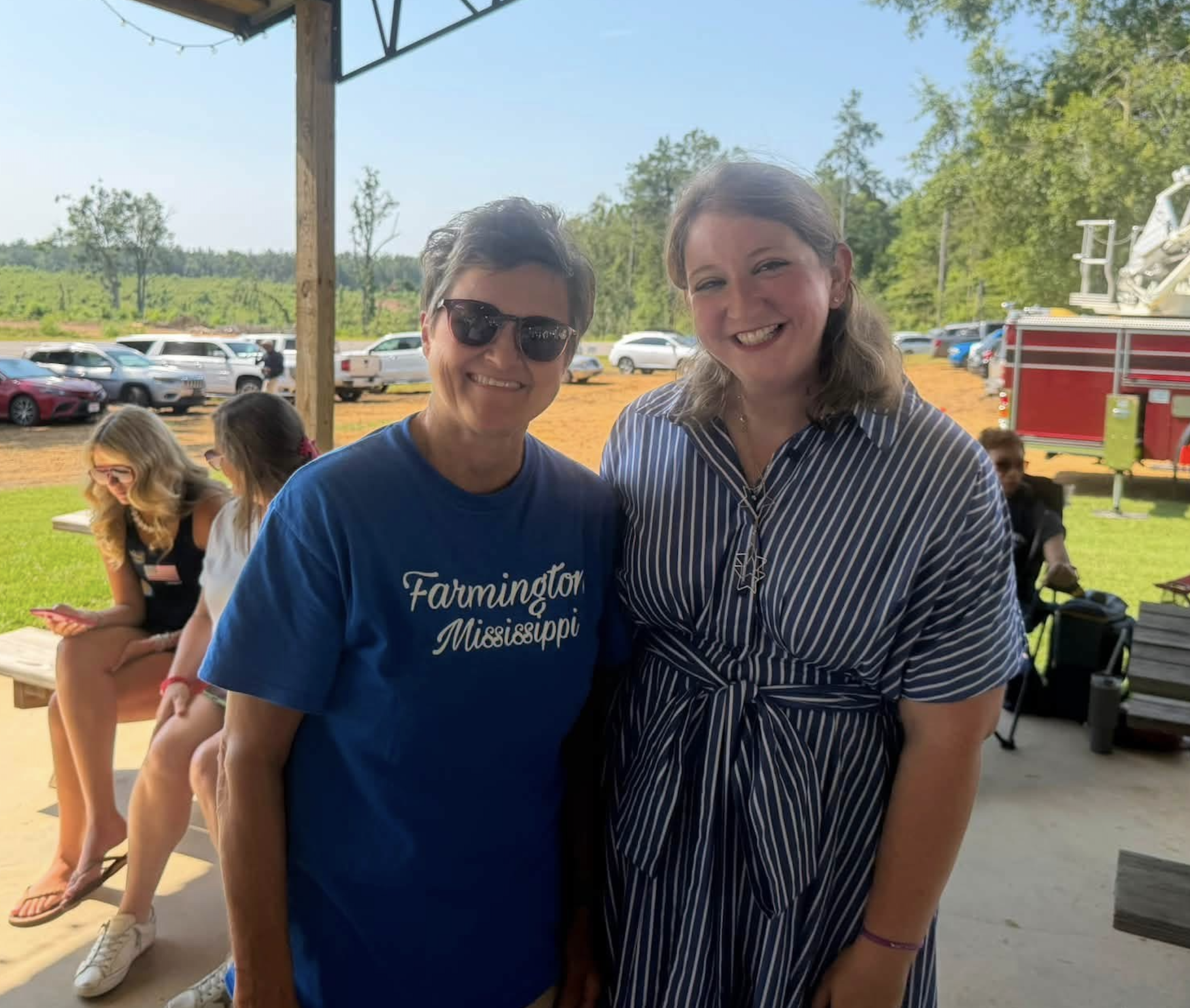 Two women smiling at an outdoor event, with other people sitting and standing in the background, and a line of cars and green trees beyond a fence.