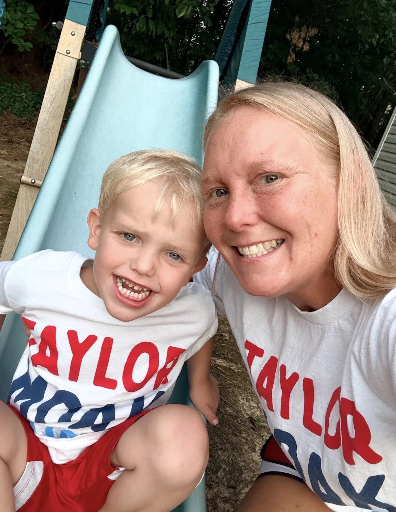 A woman and a young boy smiling and taking a selfie together outdoors near a slide.