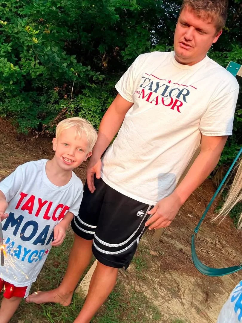 A young boy and an adult male, both wearing 'Taylor for Mayor' campaign shirts, standing outdoors in front of green bushes. The boy is smiling and holding a lit sparkler, while the man is looking down with a neutral expression. A swing set is visible