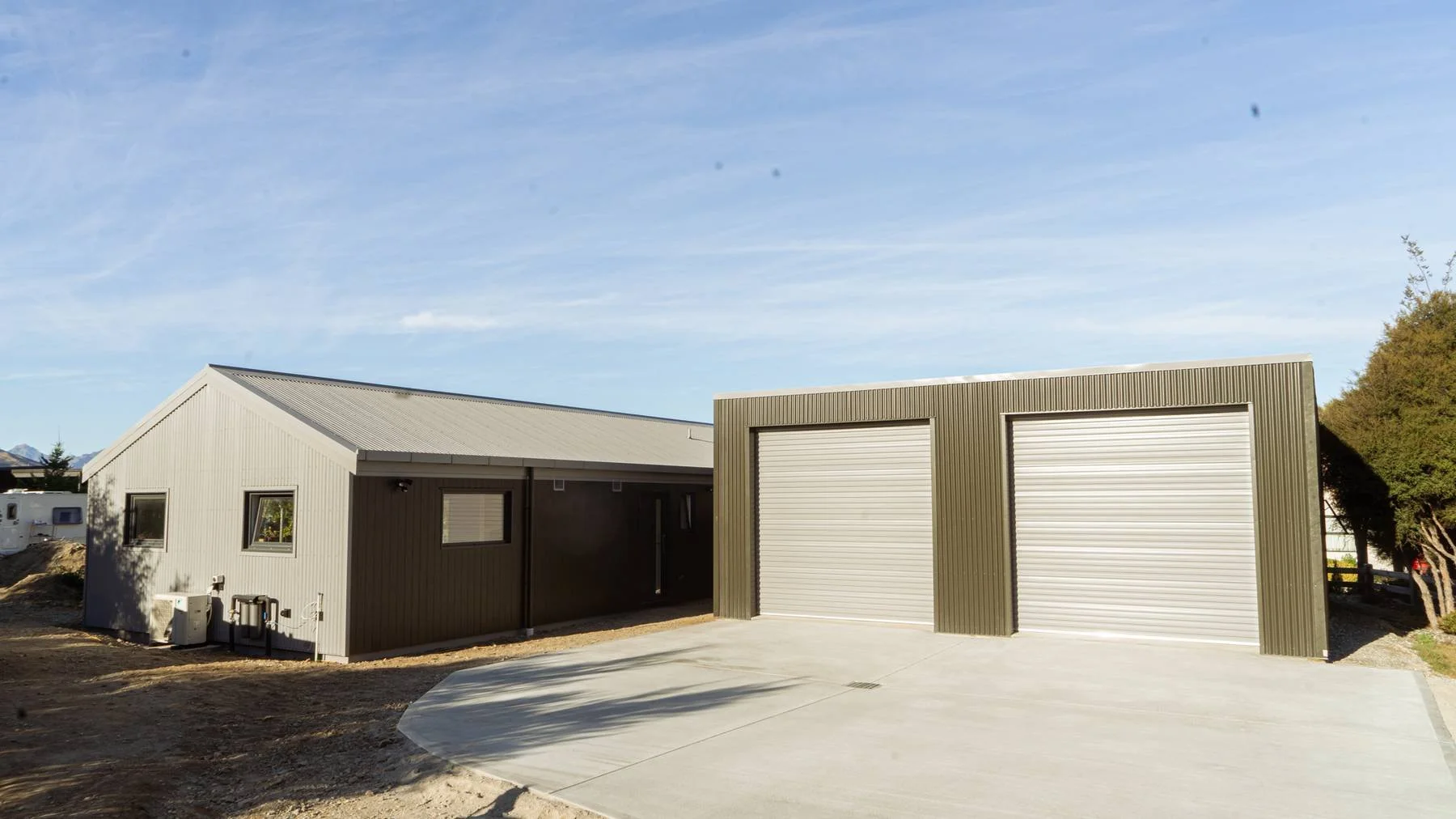 A modern house with a gable roof and beige siding, attached to a garage with two roll-up doors, set on a concrete driveway under a blue sky, with trees in the background.