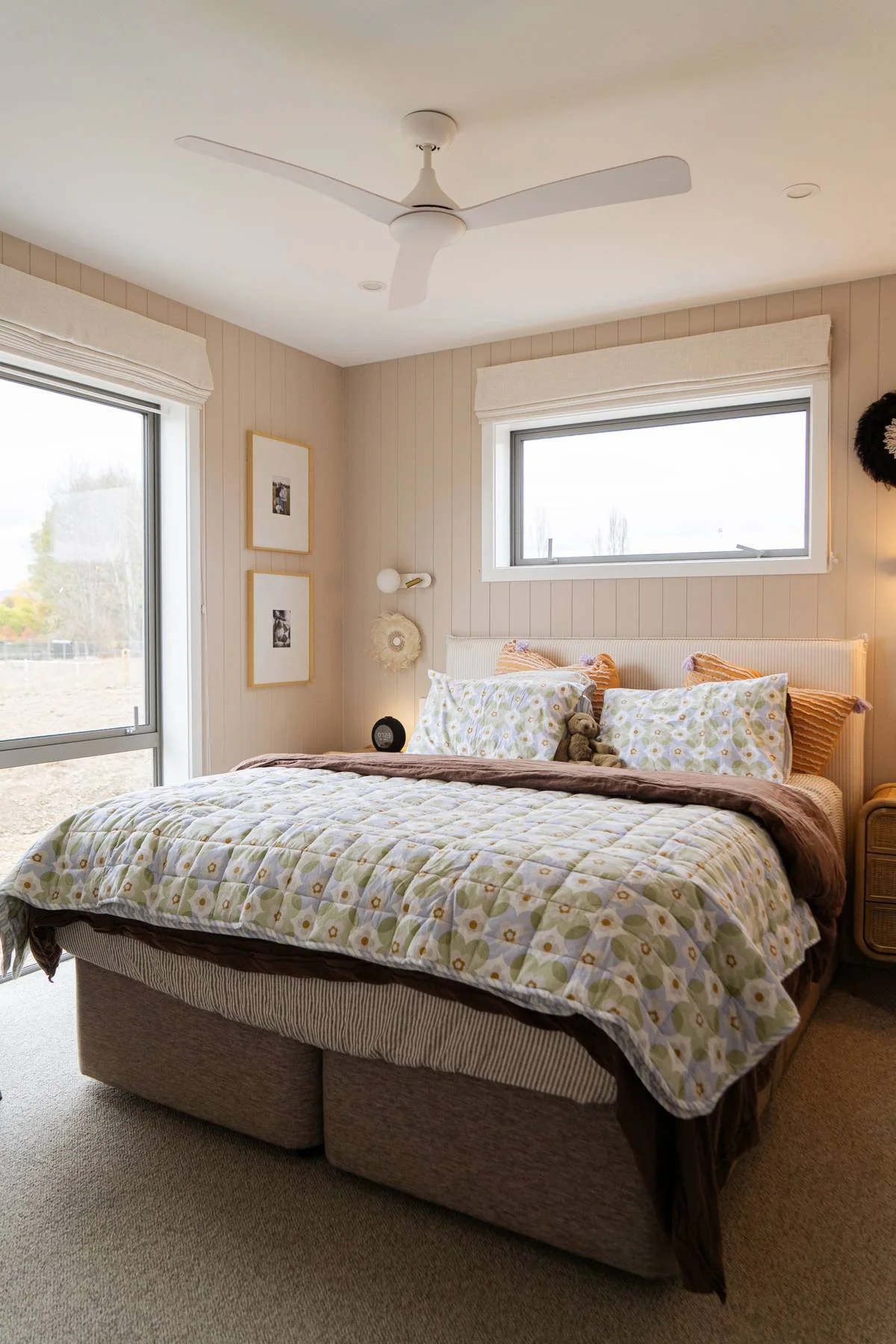 Bedroom with a bed featuring floral bedding, pillows, and stuffed animals, windows with white curtains, wall decorations, ceiling fan, and a side table.
