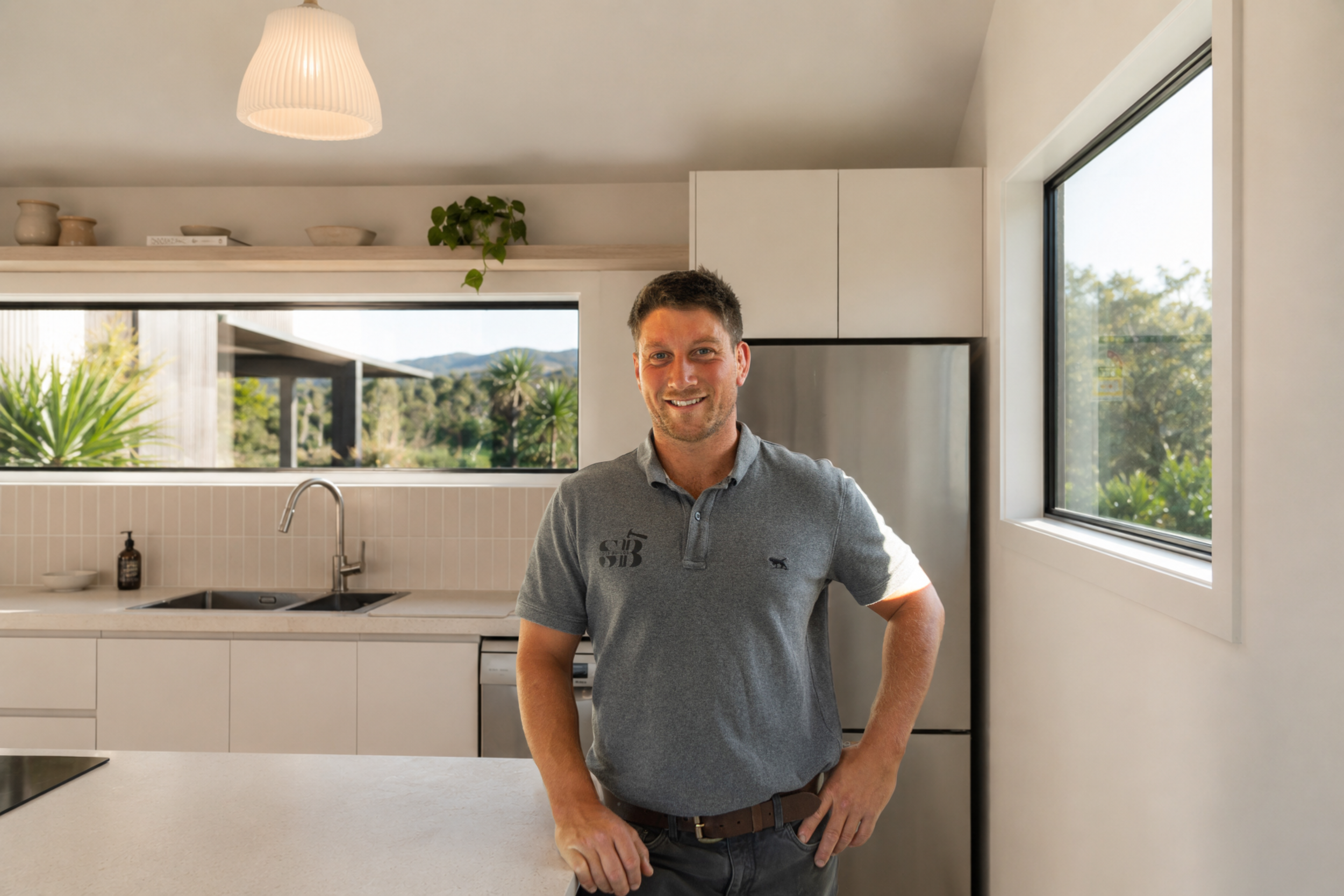 A man standing in a modern kitchen with large windows, smiling at the camera, wearing a gray polo shirt and dark pants.