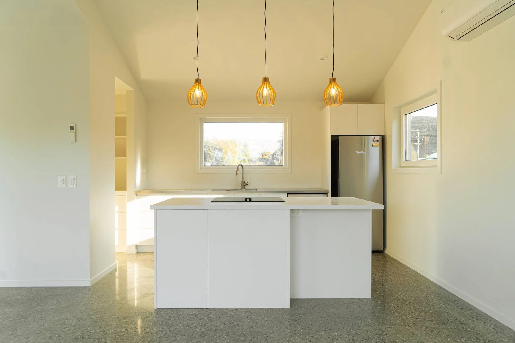 Modern kitchen with white island, natural light from windows, pendant lights hanging from ceiling, and stainless steel refrigerator.