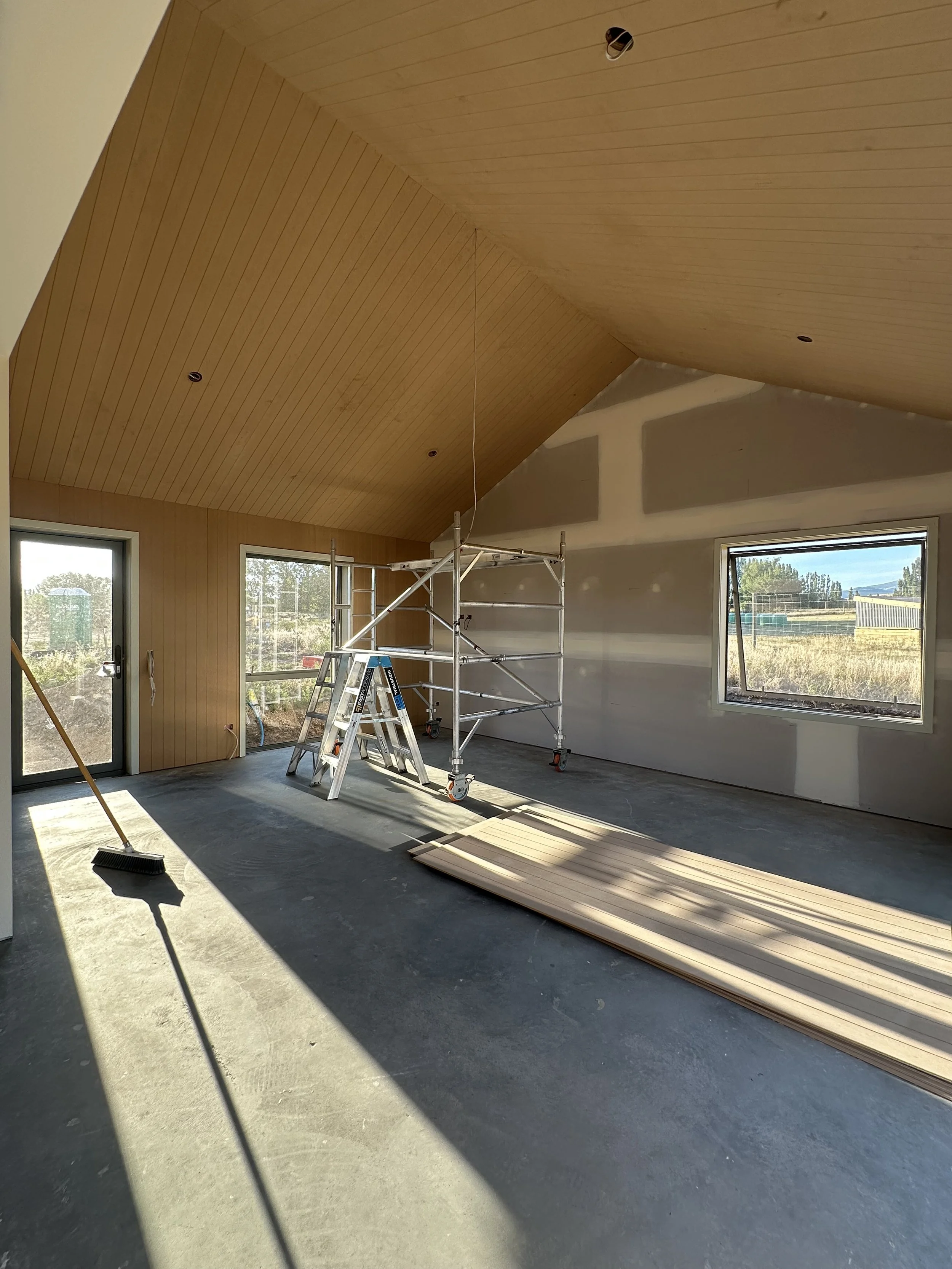 Interior room under construction with scaffolding, a ladder, wooden panels on the ceiling, and large windows showing an outdoor landscape.