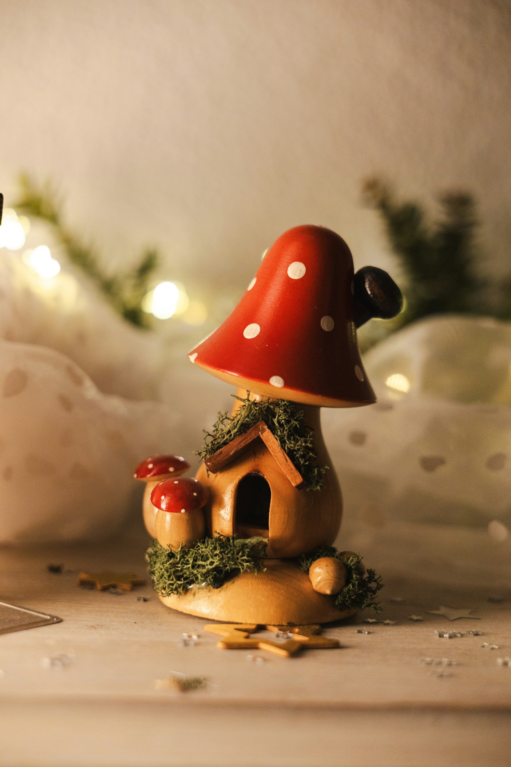 Decorative ceramic mushroom house with smaller mushrooms, moss, and fairy lights in the background, with scattered star-shaped confetti on a white surface.