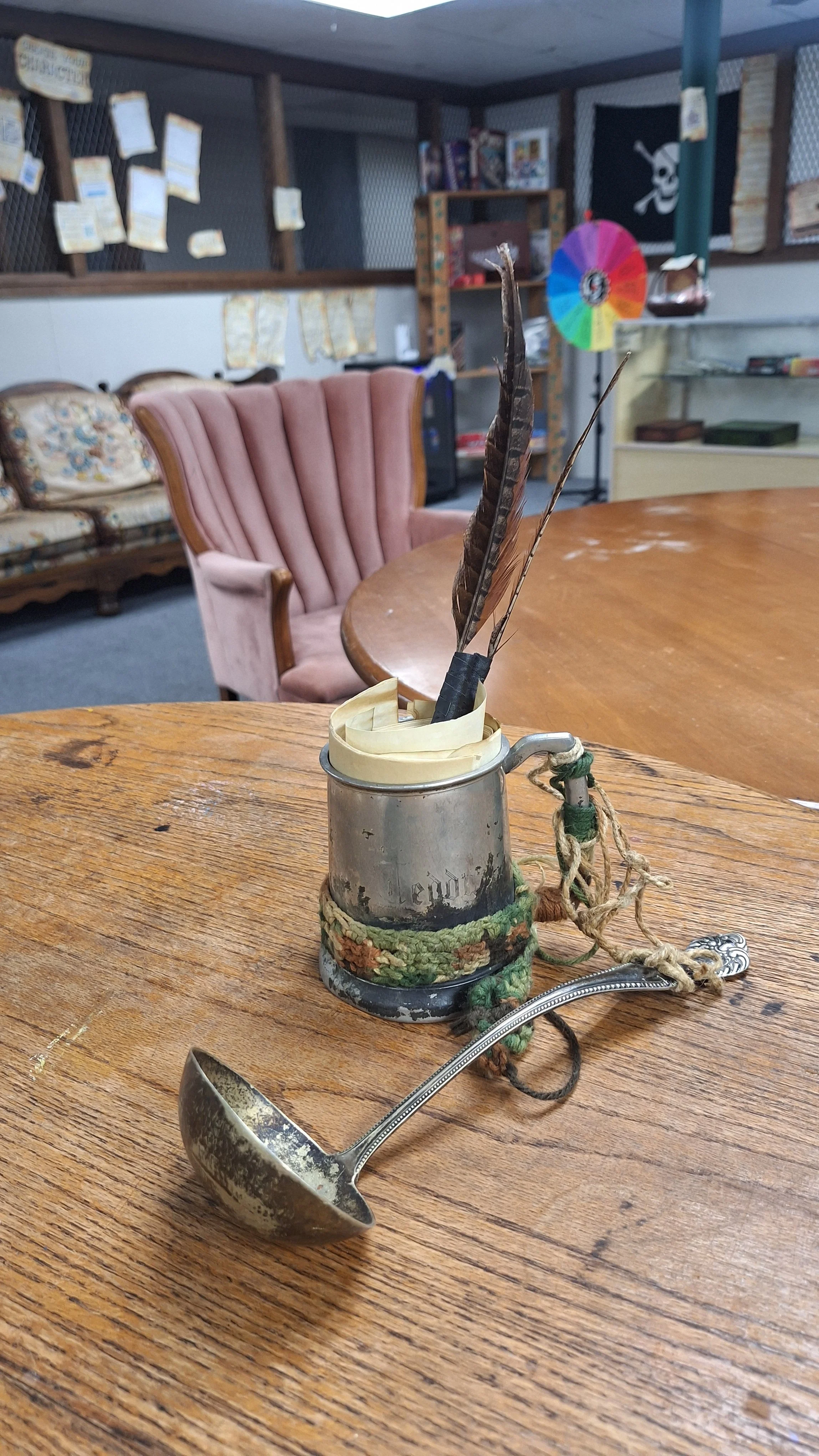 A vintage metal mug filled with feather and decorated with woven and yarn accents, placed on a wooden table. In the background, there are chairs with patterned upholstery, shelves with books, a spinning wheel, and various wall decorations including a black flag with a skull and crossbones, and a colorful spinning wheel.