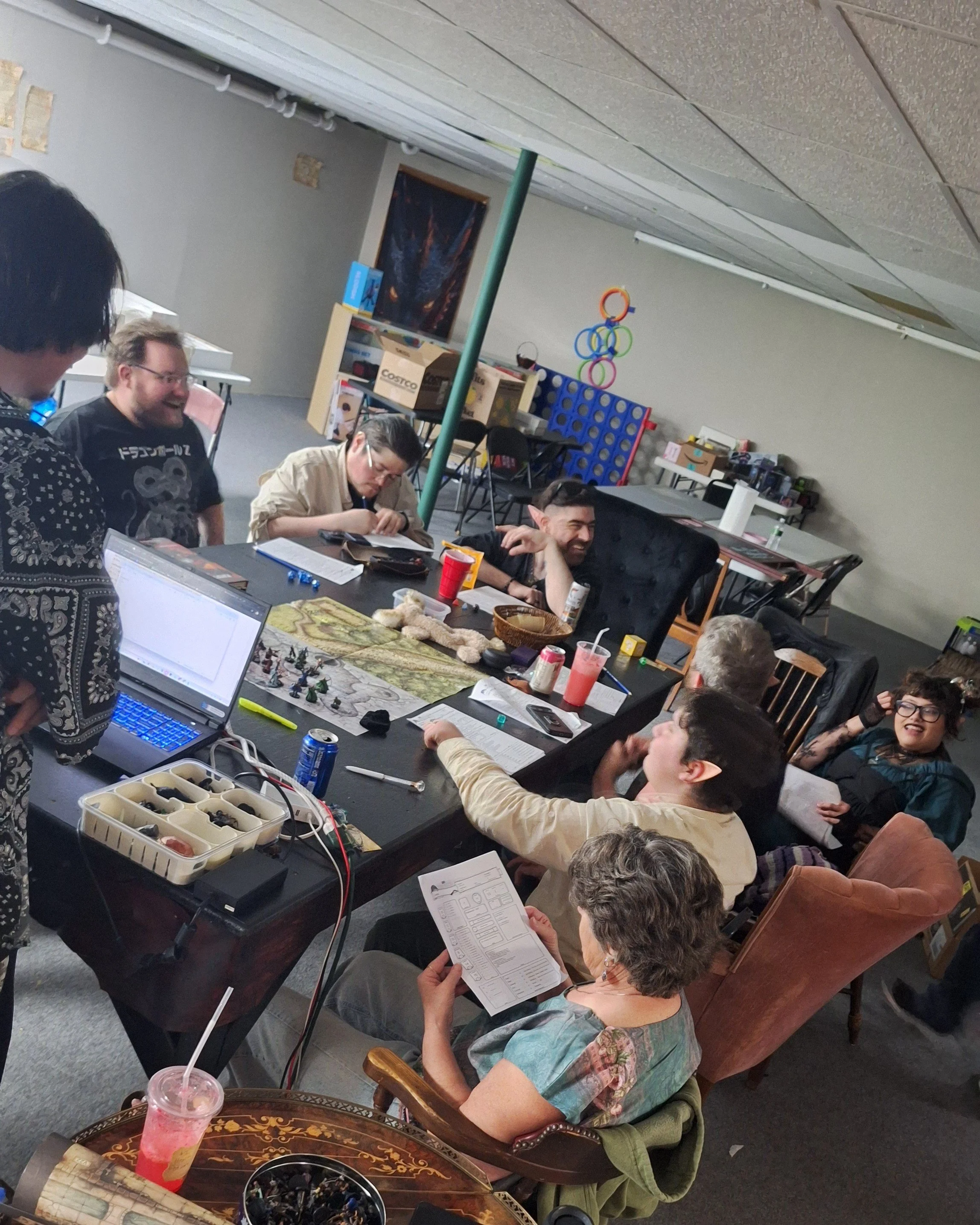 Group of people gathered around a table playing a tabletop role-playing game with papers and miniatures, in a room decorated with board game storage and a Connect Four game in the background.