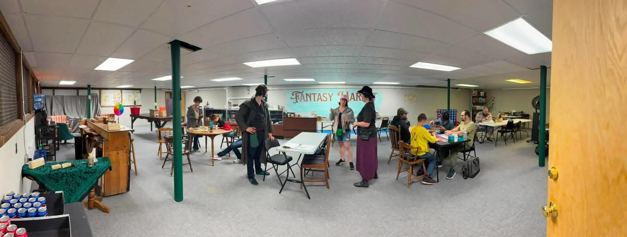 People playing a tabletop role-playing game in a community center decorated with a sign that reads 'FANTASY MARKET' and a colorful spinning wheel in the background.