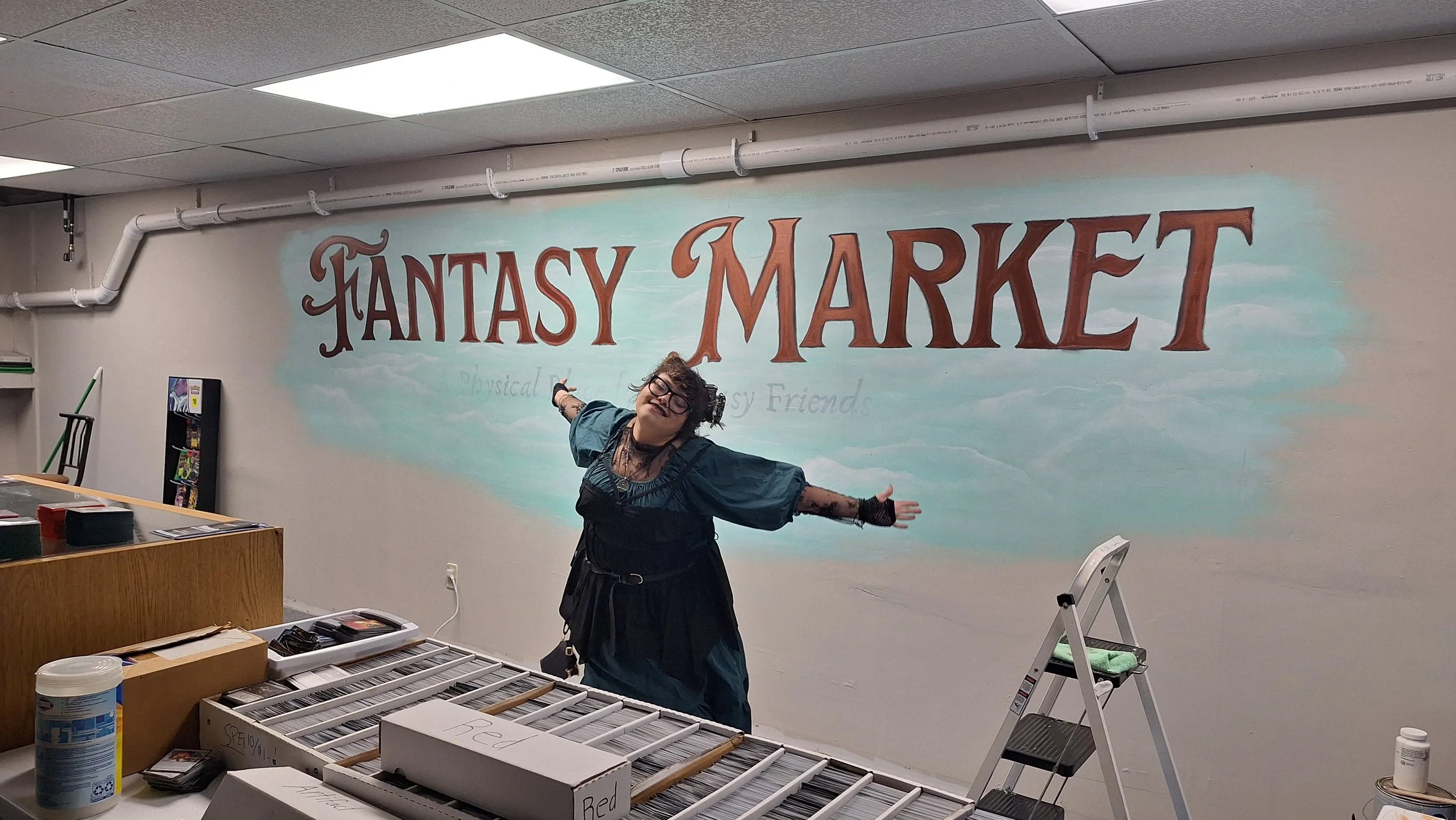 Person standing in front of a colorful mural that says 'FANTASY MARKET' in large letters, inside a room with shelves and tables.