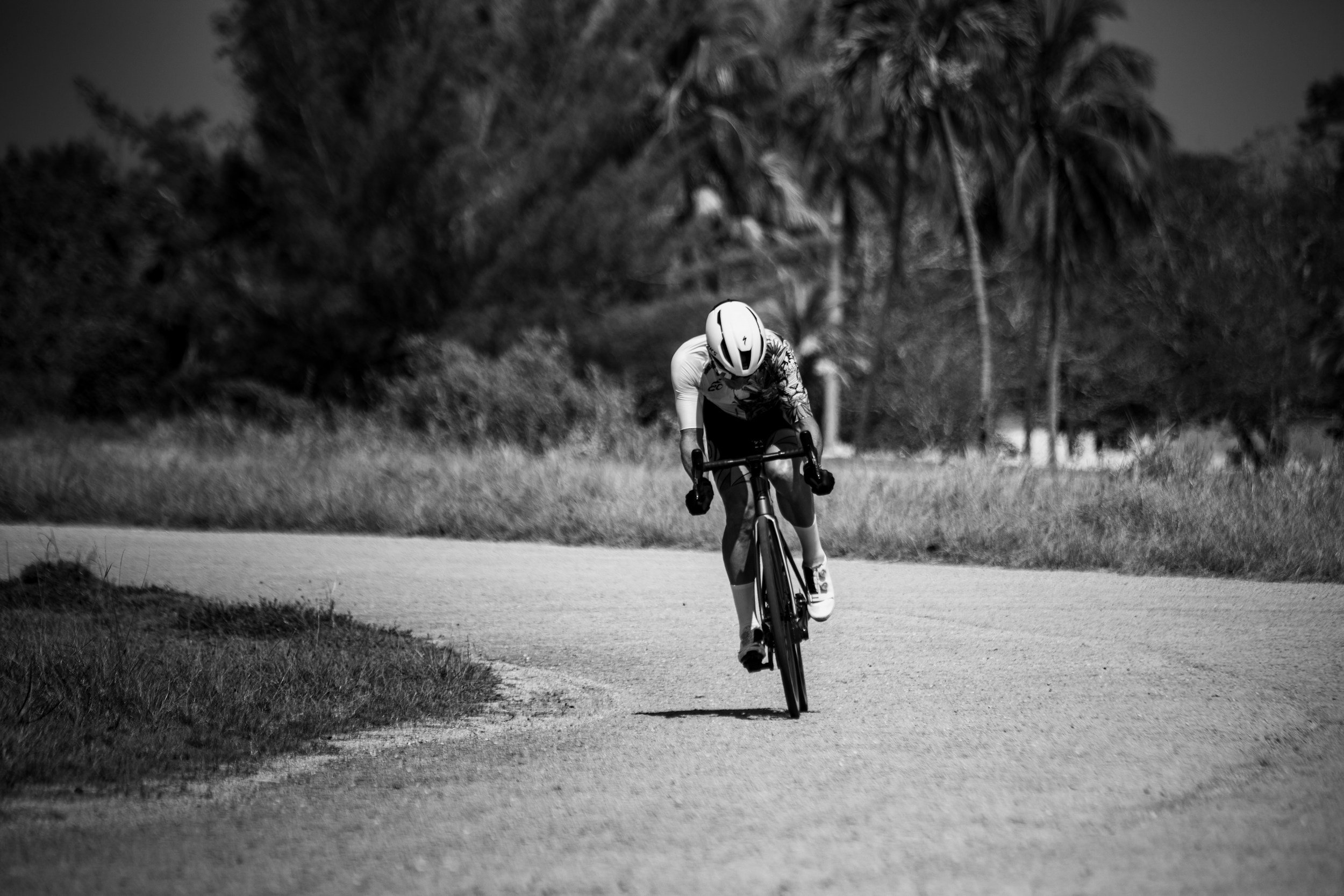 A cyclist wearing a helmet and sunglasses riding a bike along a scenic path with trees and grass in the background, in black and white.