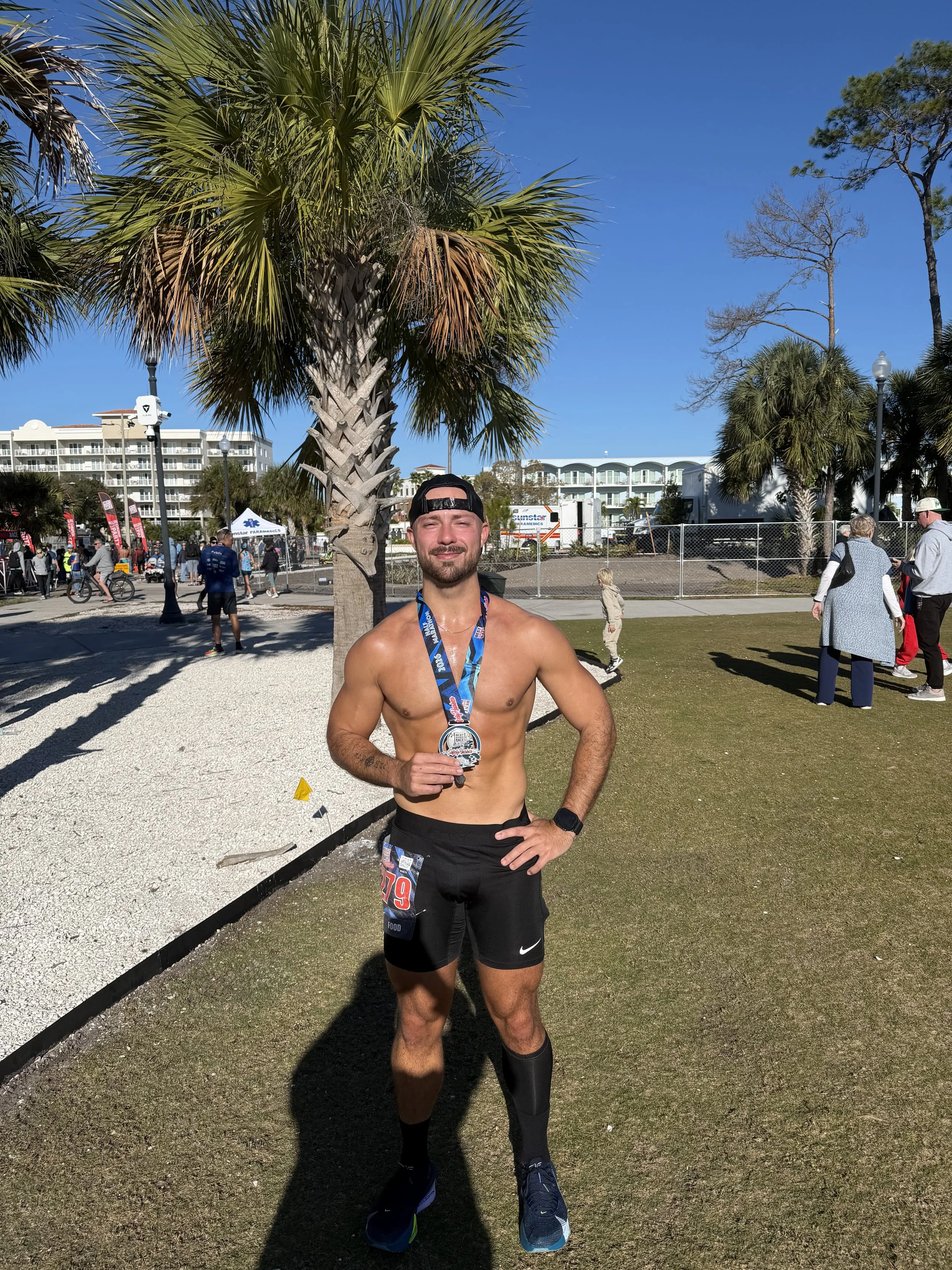 A shirtless man wearing running gear and a medal around his neck, standing outdoors in a park with palm trees and people in the background after a race.