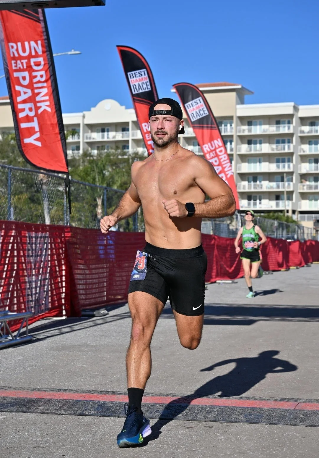 A shirtless man wearing black running shorts and a black cap participating in a race, with another runner behind him, on a sunny day near red barriers and flags that read 'Run Eat Drink Party' and 'Best Dam Race'.